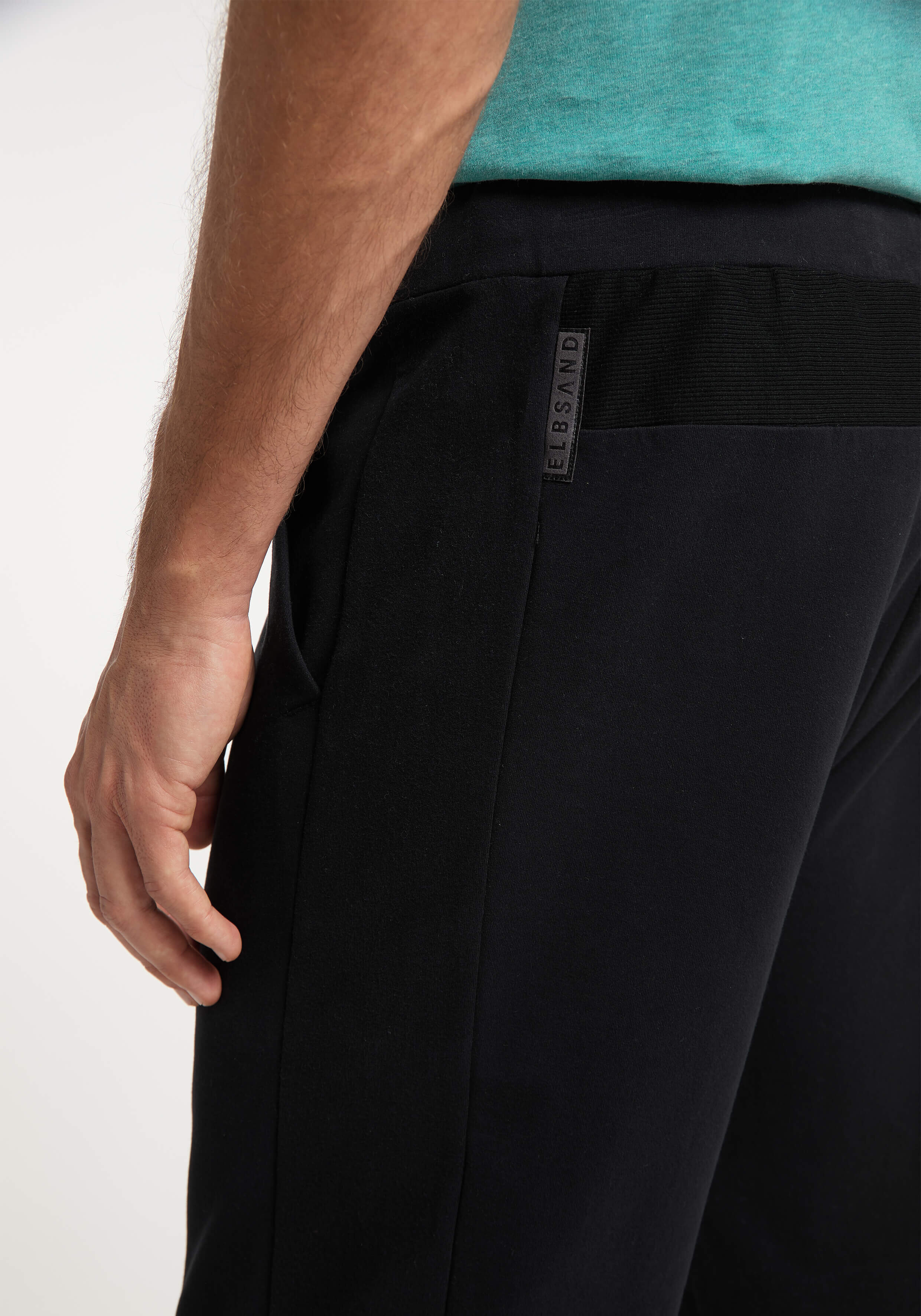 Nahaufnahme der Hand eines Mannes auf einer schwarzen Freizeithose mit ELBSAND-Label.