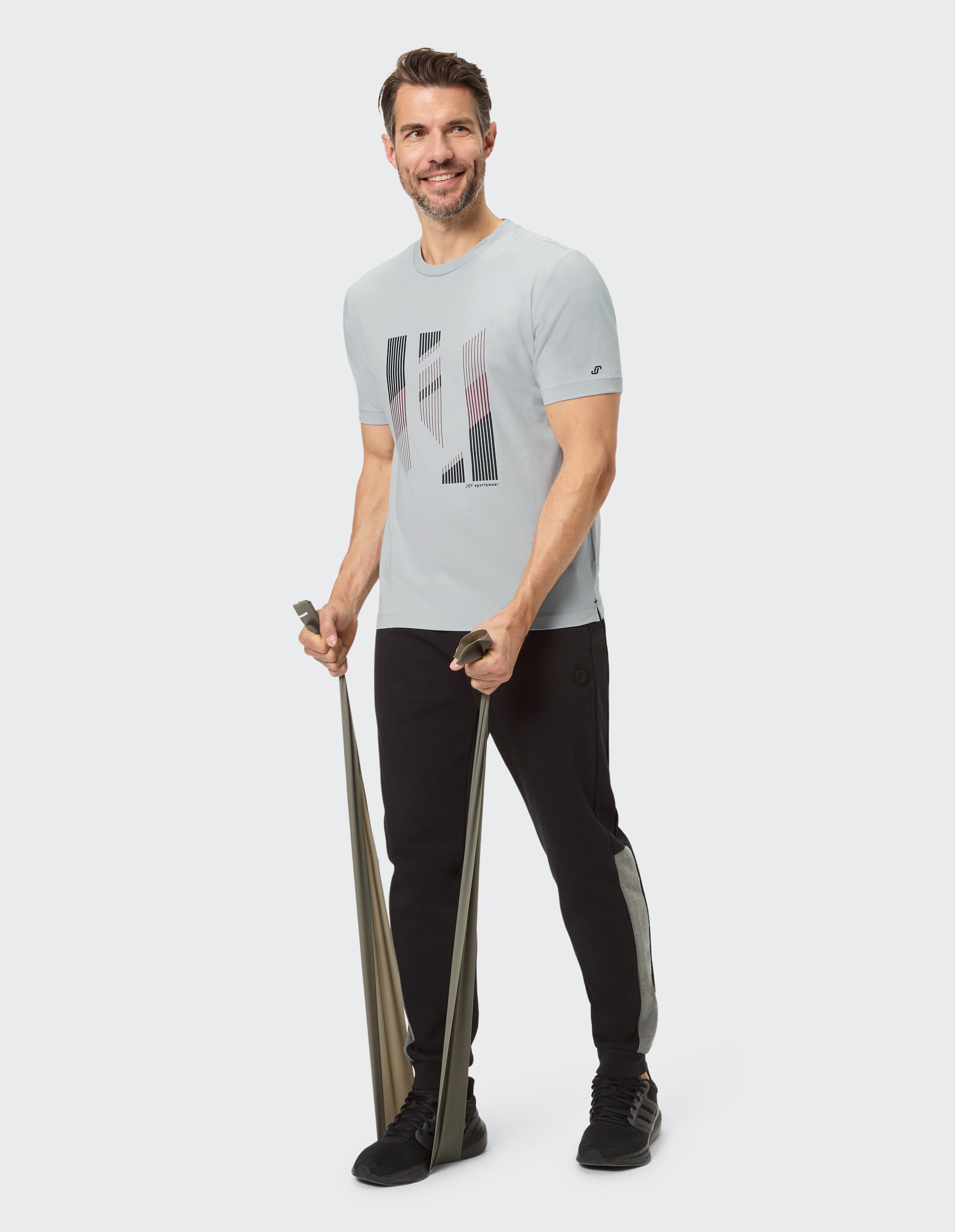 Mann in hellgrauem Grafik-T-Shirt und schwarzer Jogginghose, der Widerstandsbänder benutzt.