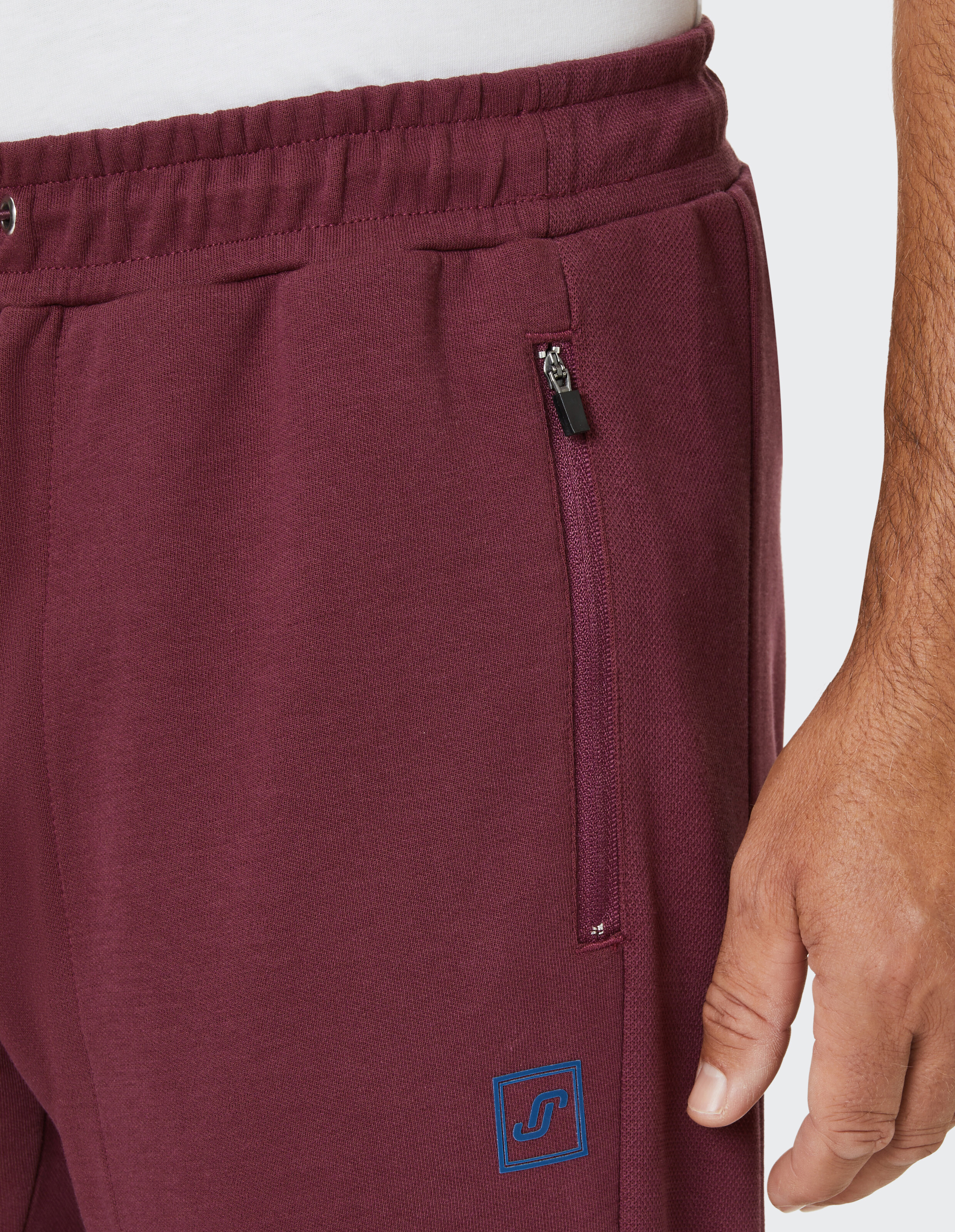 Bordeauxrote Jogginghose mit elastischem Bund, Reißverschlusstasche und blauem Logo, Hand an der Seite sichtbar.