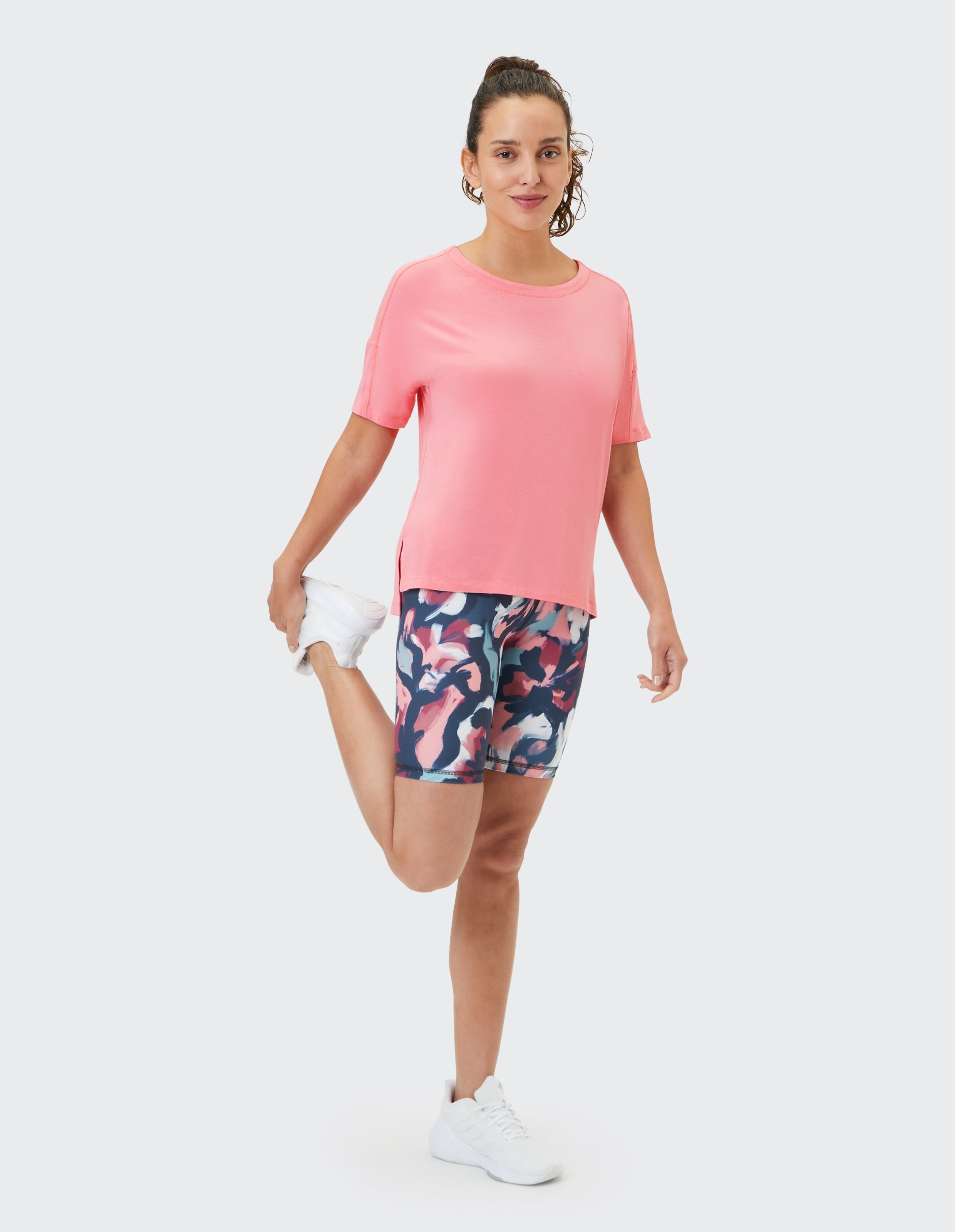 Frau in rosa T-Shirt und gemusterten Bikershorts beim Stretching in lässiger Sportkleidung.