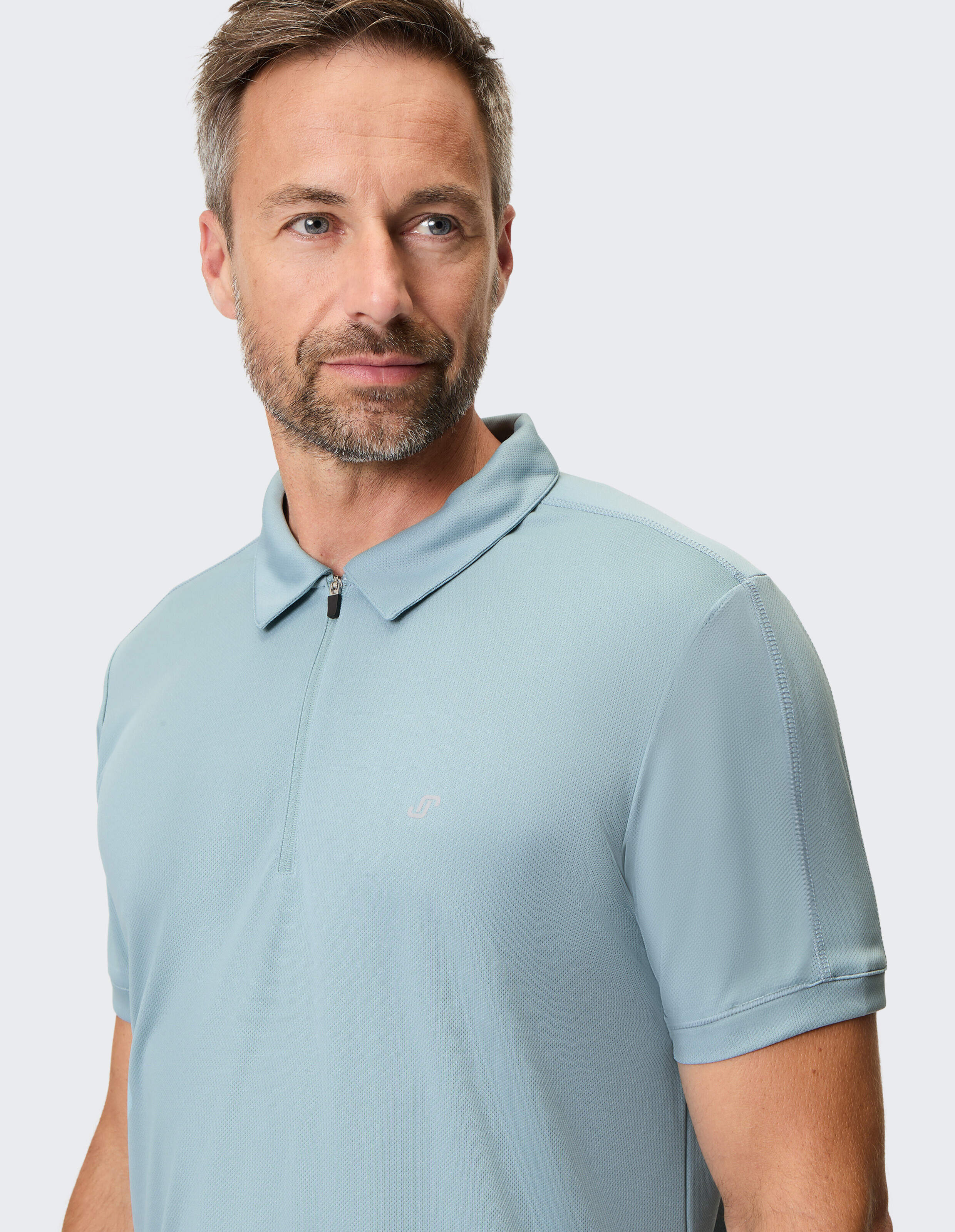 Ein Mann mit kurzem Haar und Bart trägt ein hellblaues Poloshirt mit Zipper vor neutralem Hintergrund und schaut leicht zur Seite.