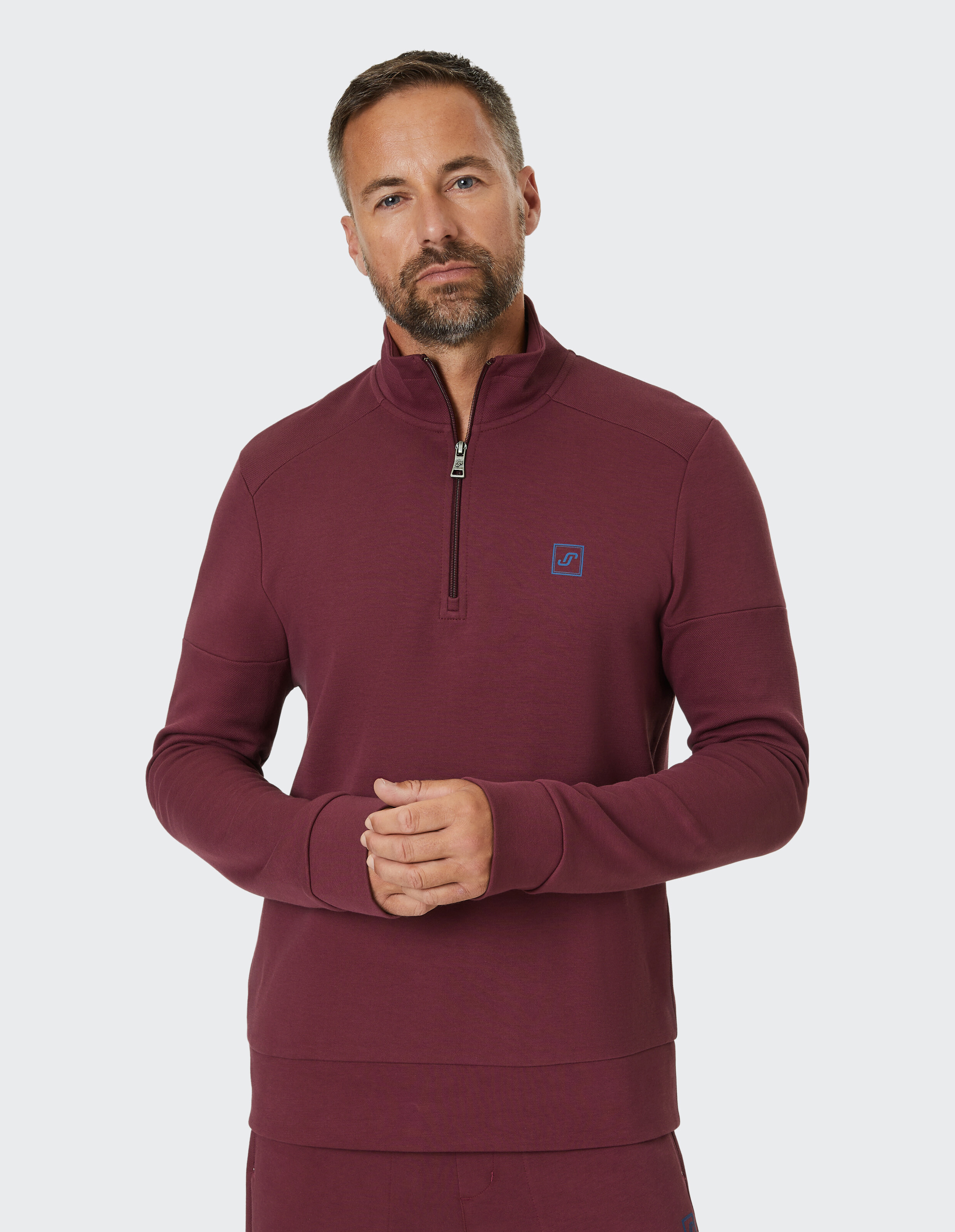 Ein bordeauxroter Pullover mit Stehkragen und kurzem Reißverschluss, verziert mit einem kleinen blauen Logo auf der Brust.