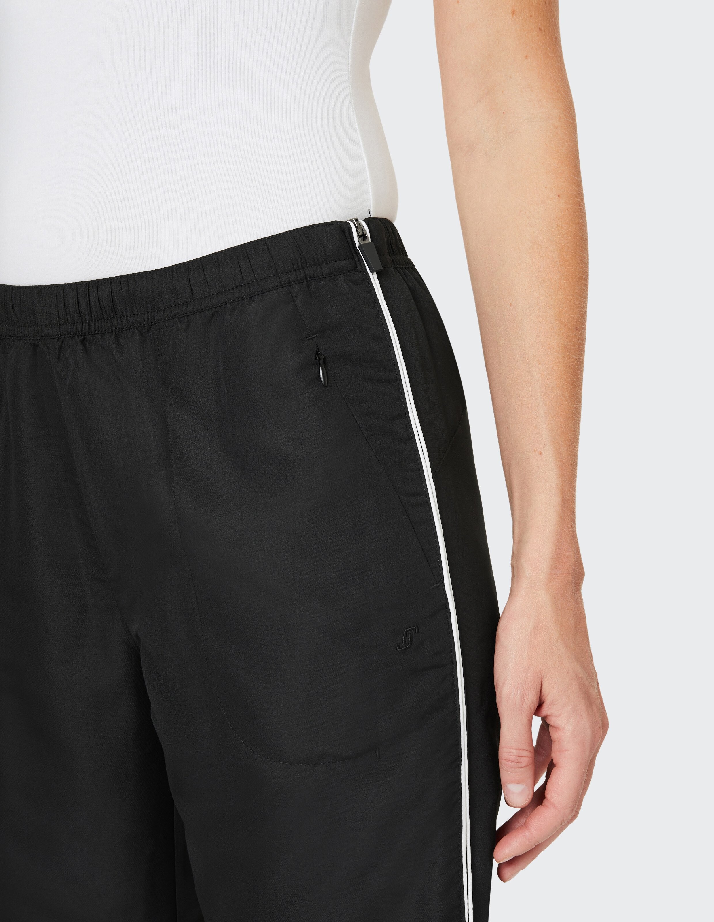 Person trägt ein weißes Oberteil und eine schwarze Hose mit elastischem Bund und seitlichem weißen Streifen, Hand locker am Bein.
