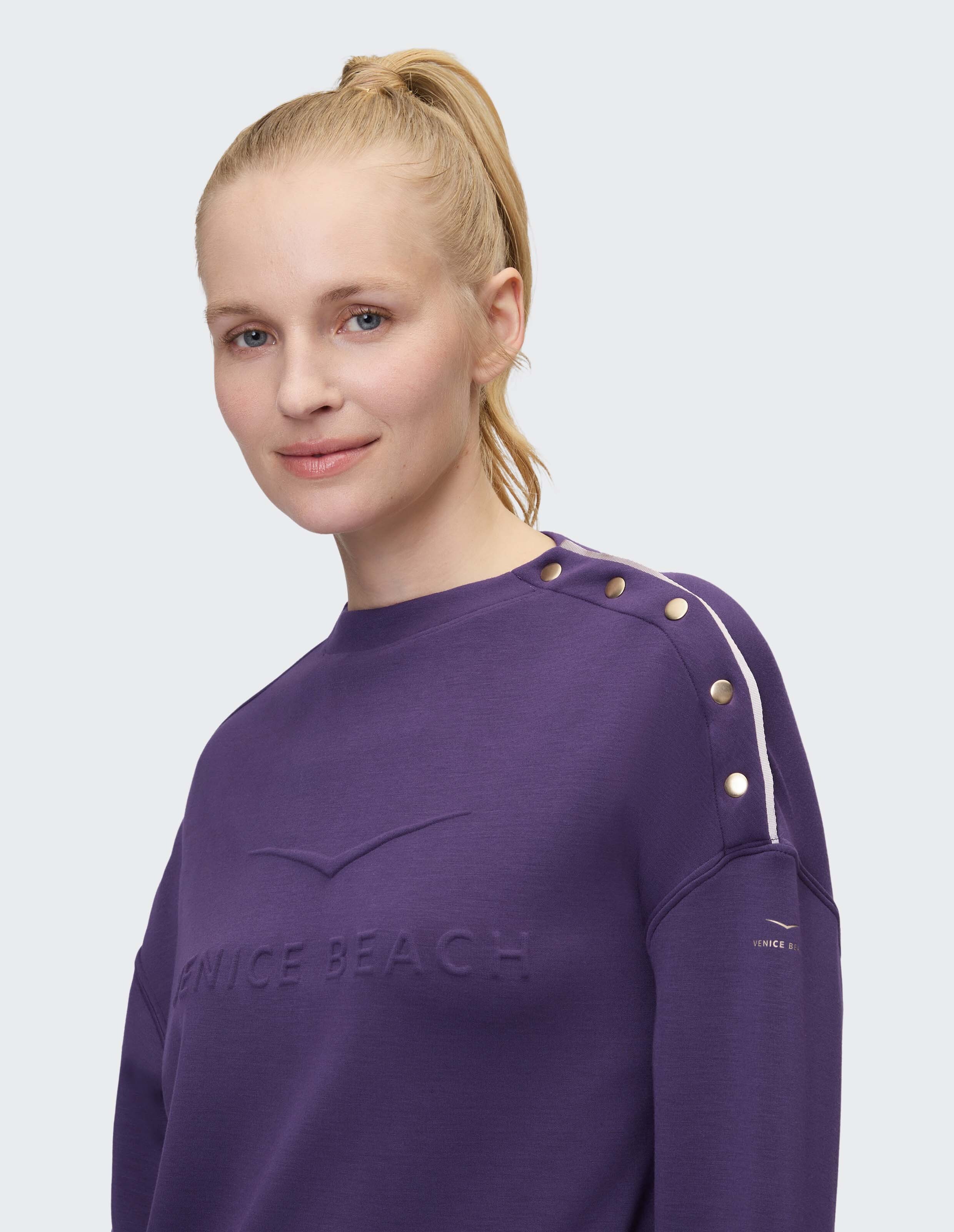 Frau trägt ein gemütliches lilafarbenes Venice Beach-Sweatshirt mit dekorativen goldenen Schulterknöpfen.