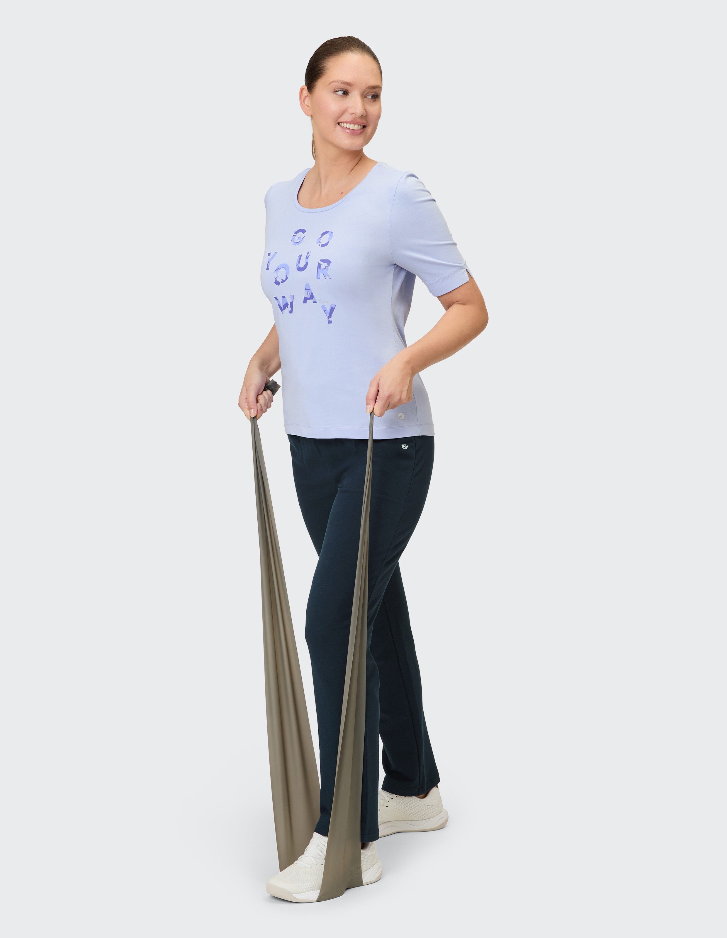Frau in einem hellvioletten T-Shirt und einer dunkelblauen Hose, hält Trainingsbänder in der Hand.