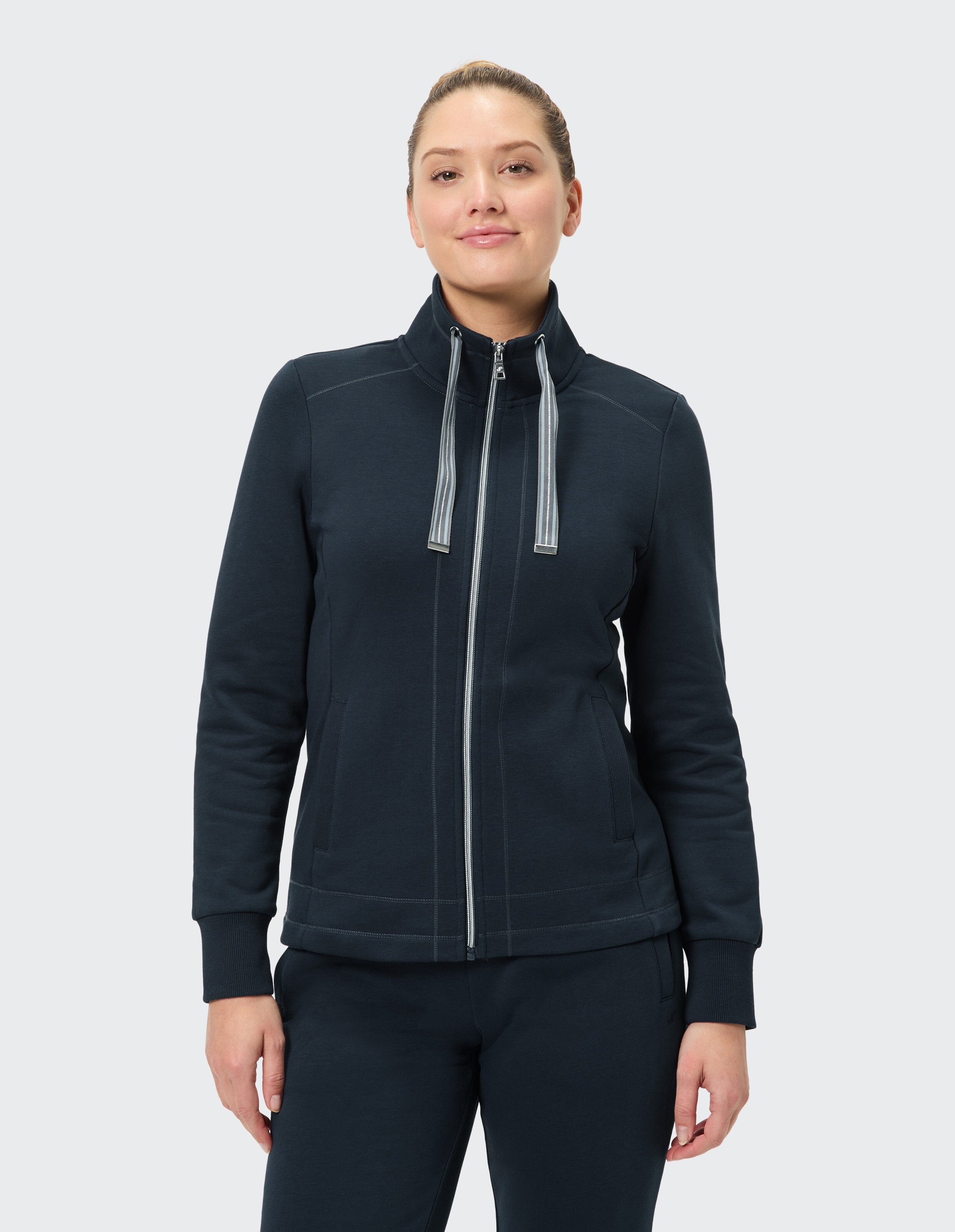 Eine Person trägt eine dunkelblaue, langärmelige Jacke mit Reißverschluss und Taschen, stehend vor einem neutralen Hintergrund.