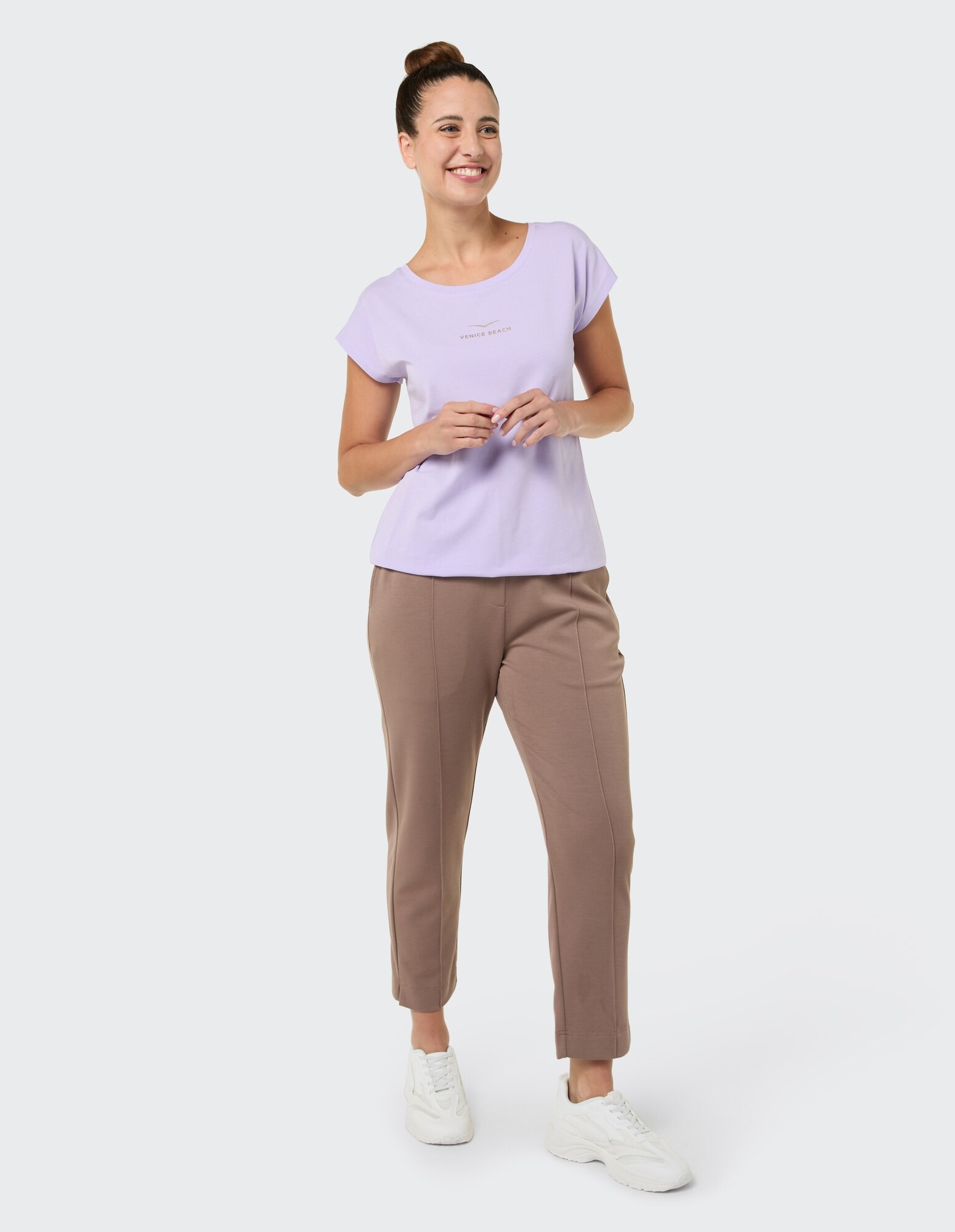 Frau in lavendelfarbenem T-Shirt und beiger Hose lächelt lässig in entspannter Atmosphäre.