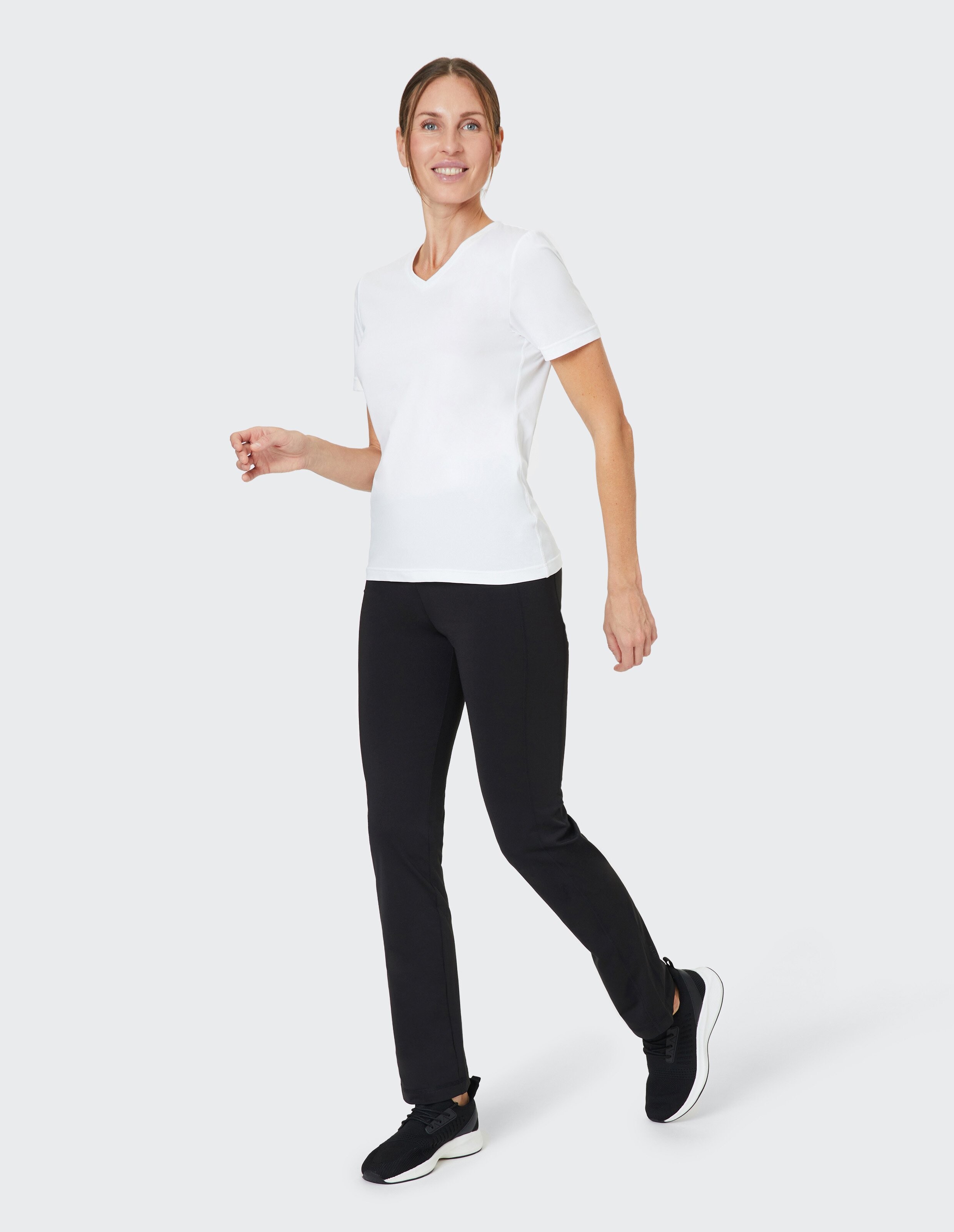 Eine Frau in einem weißen T-Shirt und schwarzen Hosen läuft lächelnd vor einem hellgrauen Hintergrund.