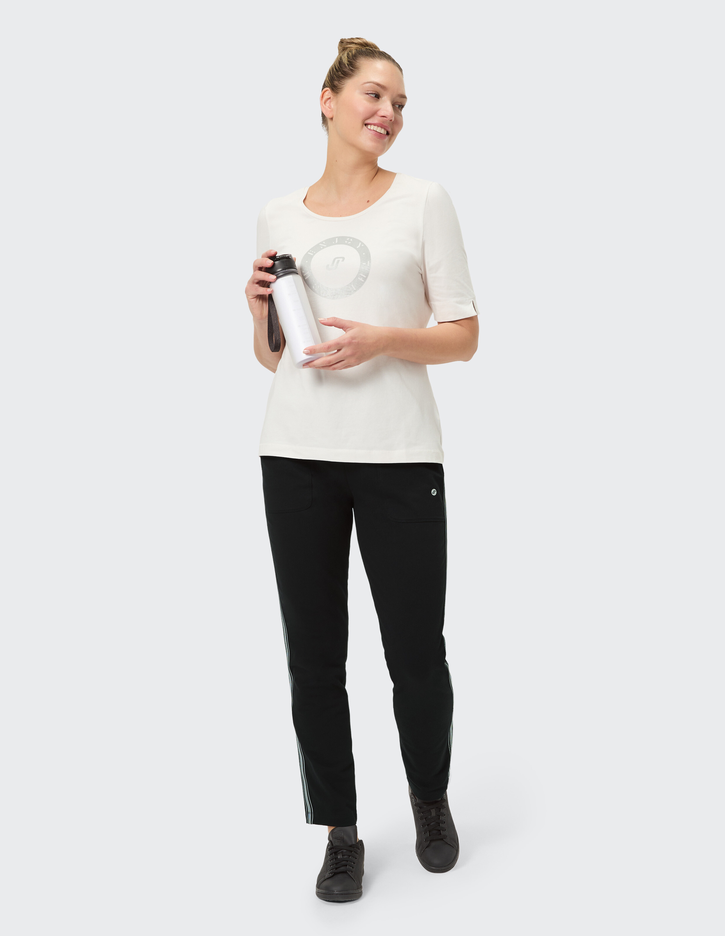 Eine Frau mit blondem Dutt und einem lächelnden Gesicht hält eine weiße Wasserflasche und trägt eine cremefarbene T-Shirt und schwarze Hose.