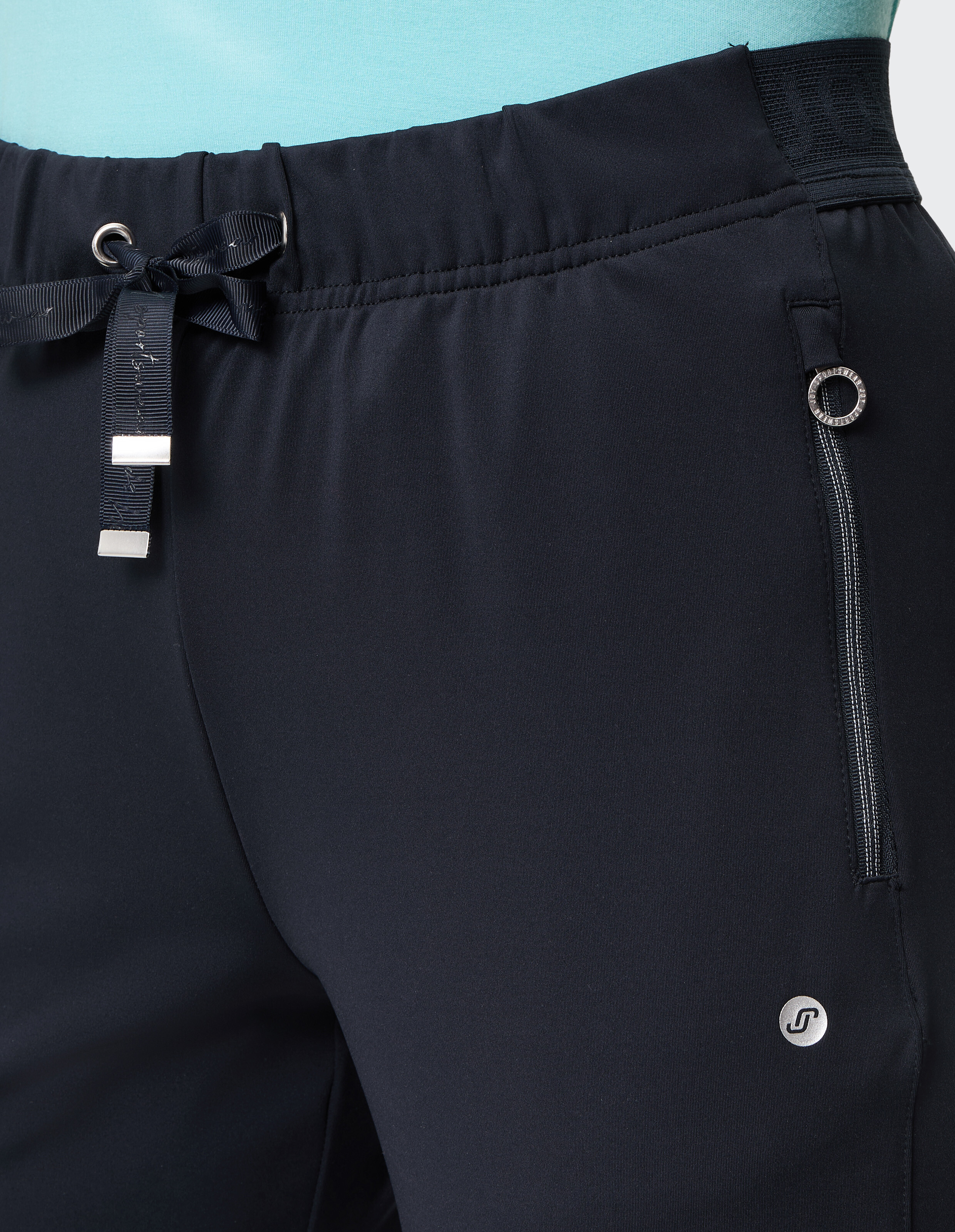 Dunkle Sporthose mit elastischem Bund und Schleifenband, seitlicher Reißverschluss und kleinem Logo, getragen über einem hellblauen Shirt.