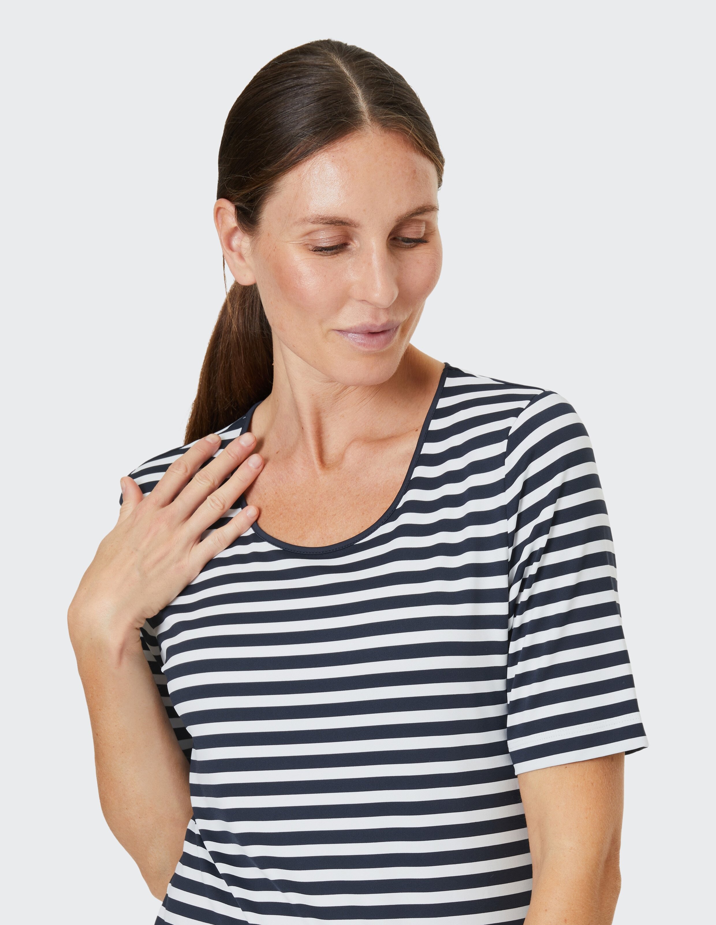 Frau mit Pferdeschwanz trägt gestreiftes Shirt, schaut nach unten und berührt sanft ihren Hals, vor hellem Hintergrund.