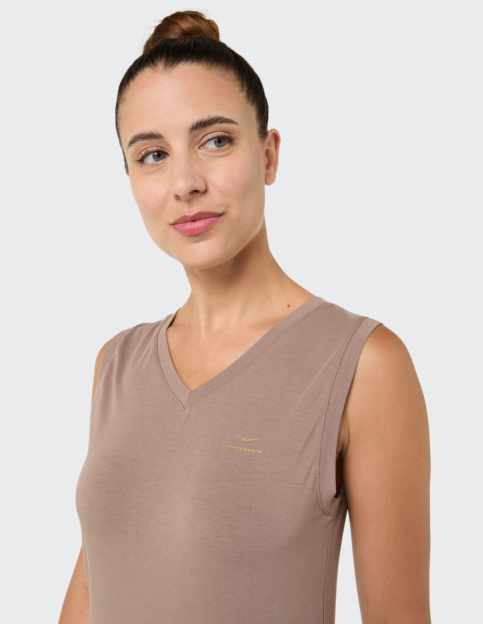 Frau in einem ärmellosen, beigefarbenen Top mit V-Ausschnitt und dezentem Logo, das einen lässigen Stil unterstreicht.
