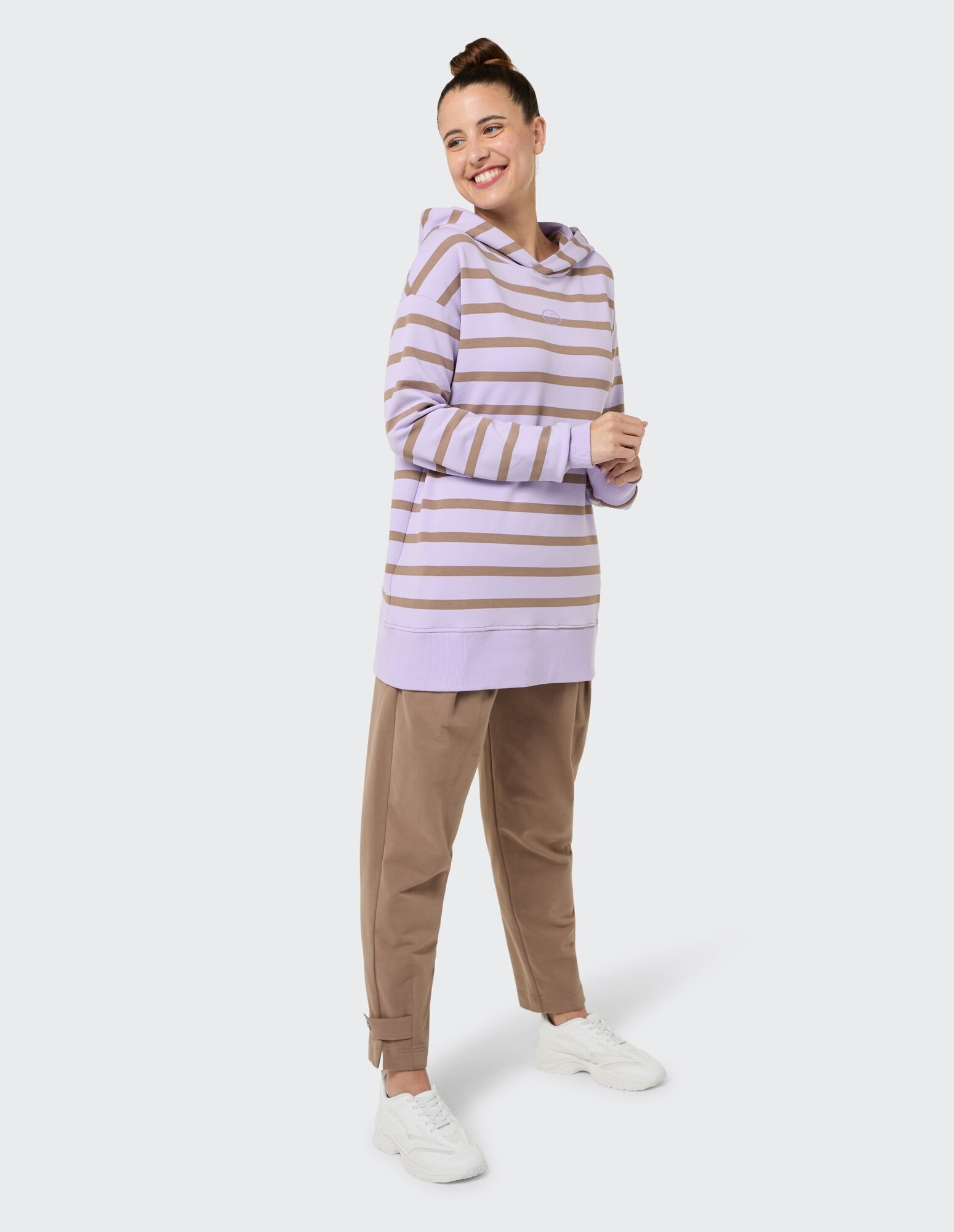 Frau in einem gestreiften lavendelfarbenen Kapuzenpullover und braunen Jogginghosen, lächelnd.