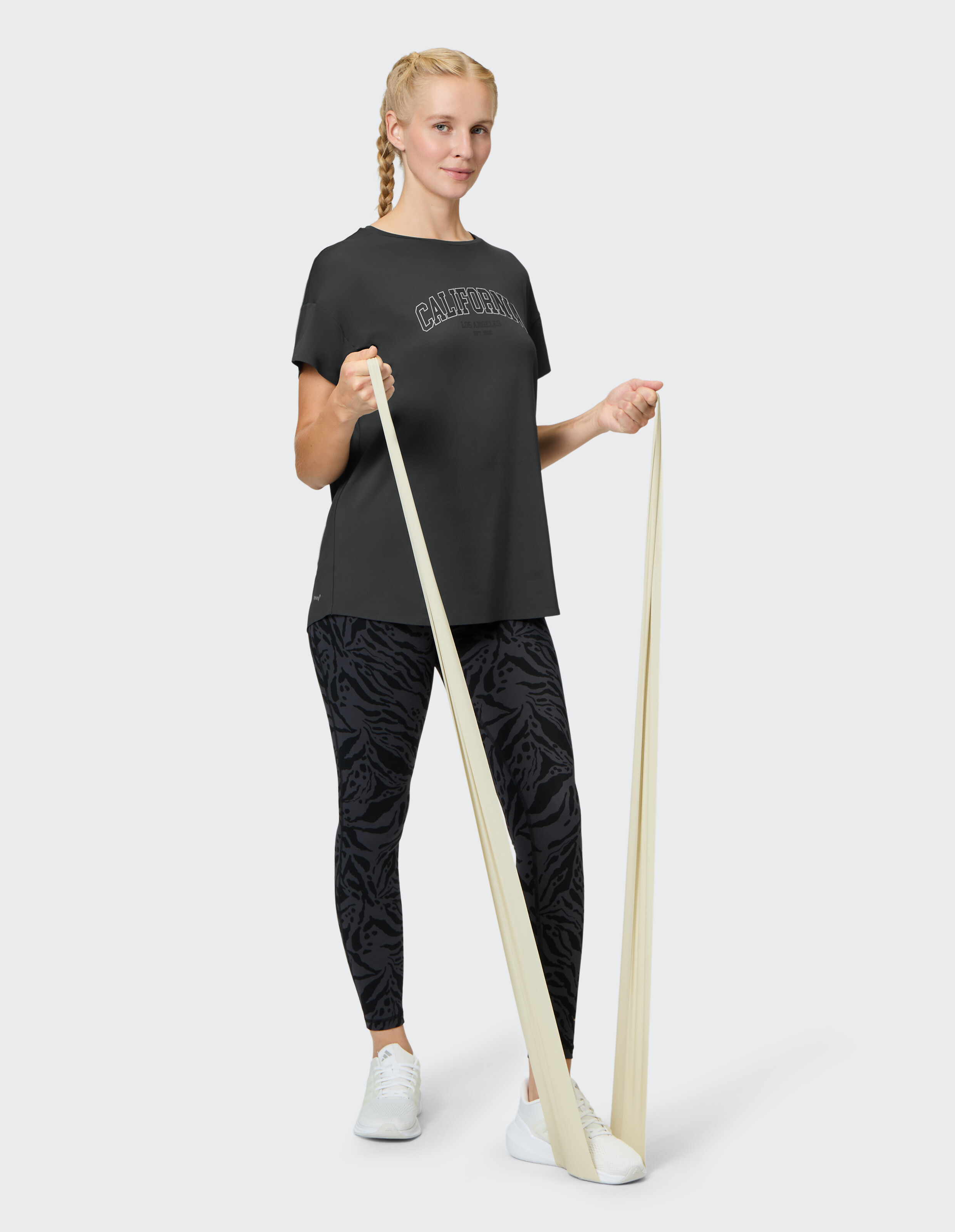 Frau in einem schwarzen California-T-Shirt und gemusterten Leggings trainiert mit einem Widerstandsband.