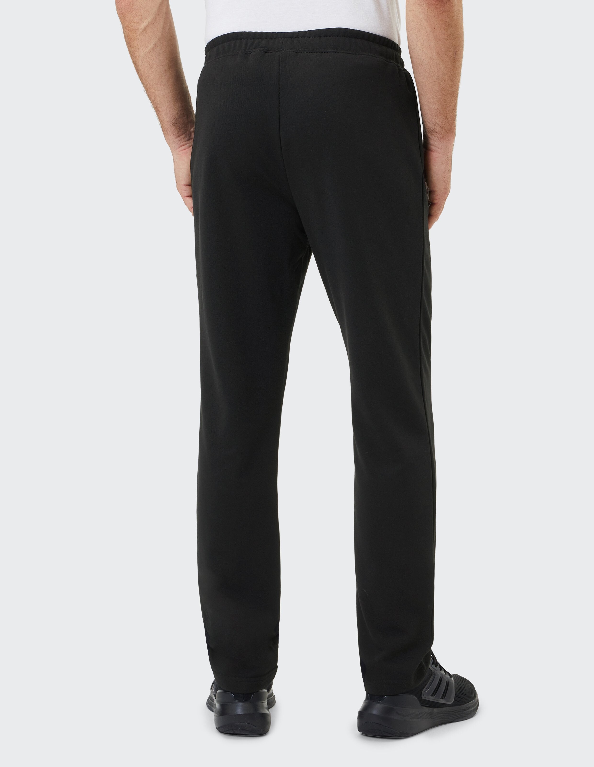 Ein Mann trägt eine schwarze Jogginghose und schwarze Sneaker, steht mit dem Rücken zur Kamera; er trägt ein weißes T-Shirt.