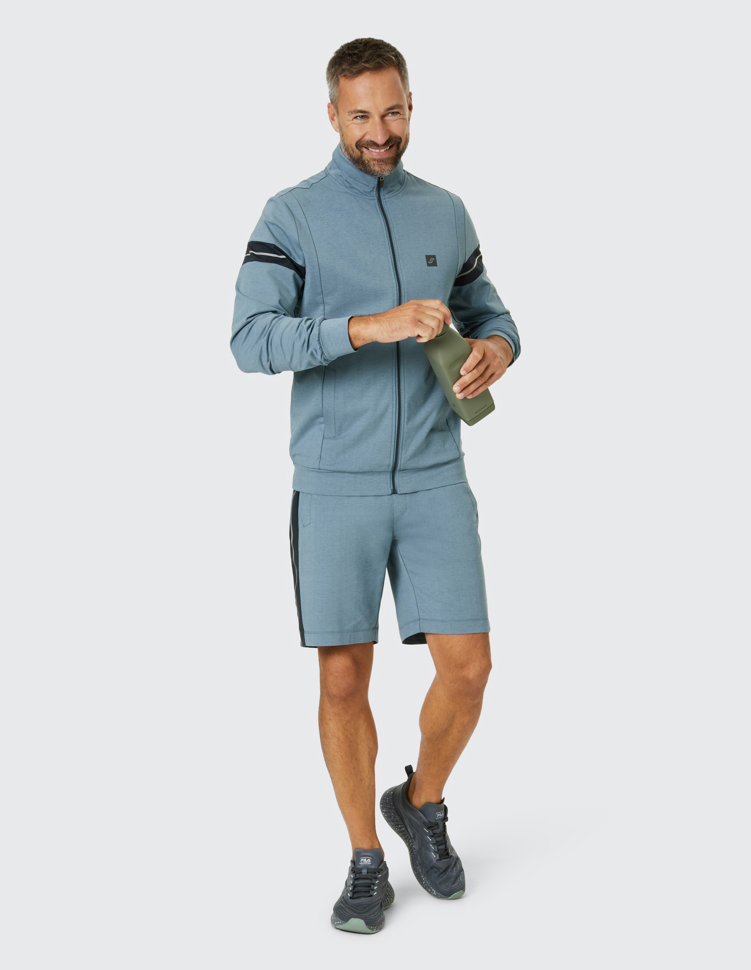 Mann in blauem Sportanzug und Sneakers hält eine grüne Wasserflasche und lächelt, während er auf hellem Hintergrund steht.