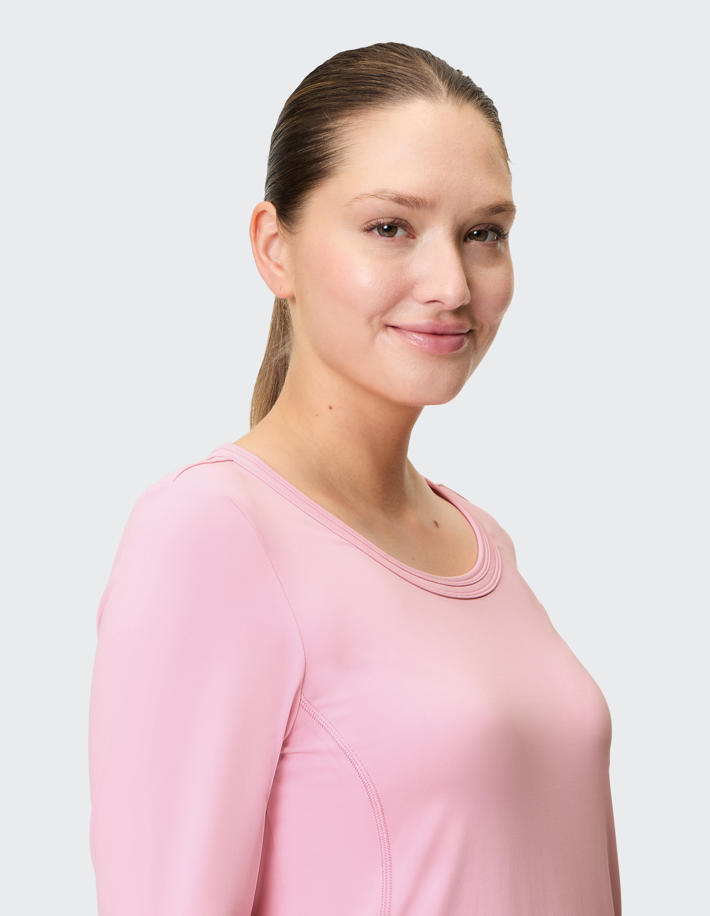 Eine lächelnde Person mit zurückgebundenem Haar trägt ein rosafarbenes Shirt vor hellem Hintergrund.