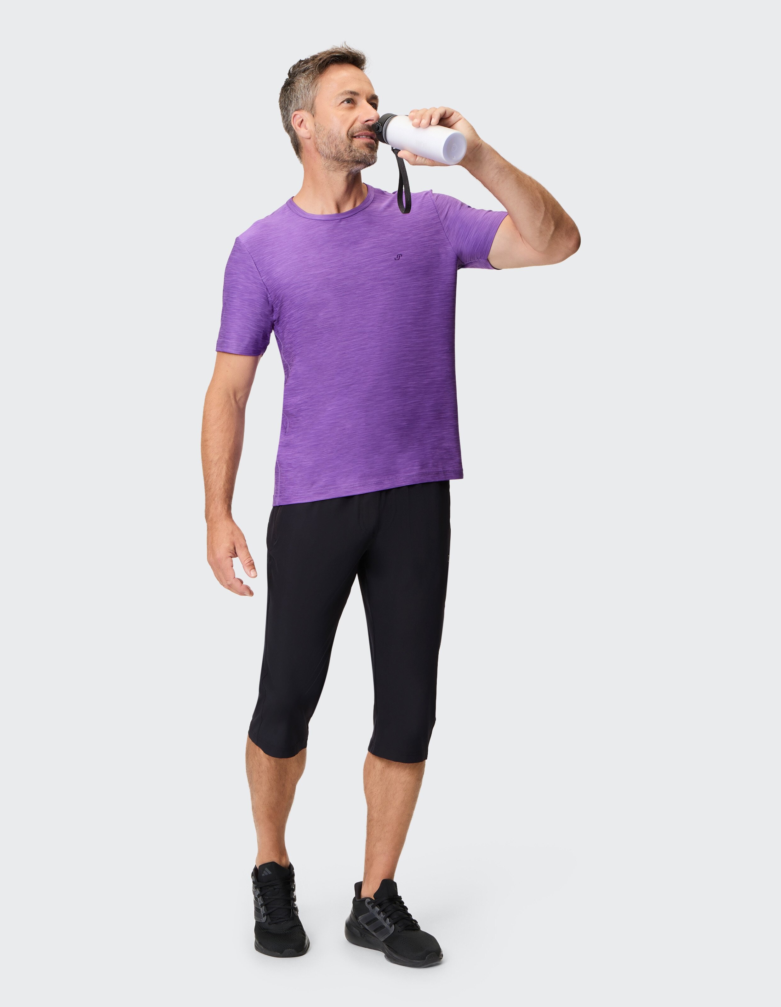 Mann in lila T-Shirt und schwarzer Caprihose trinkt aus einer Wasserflasche.