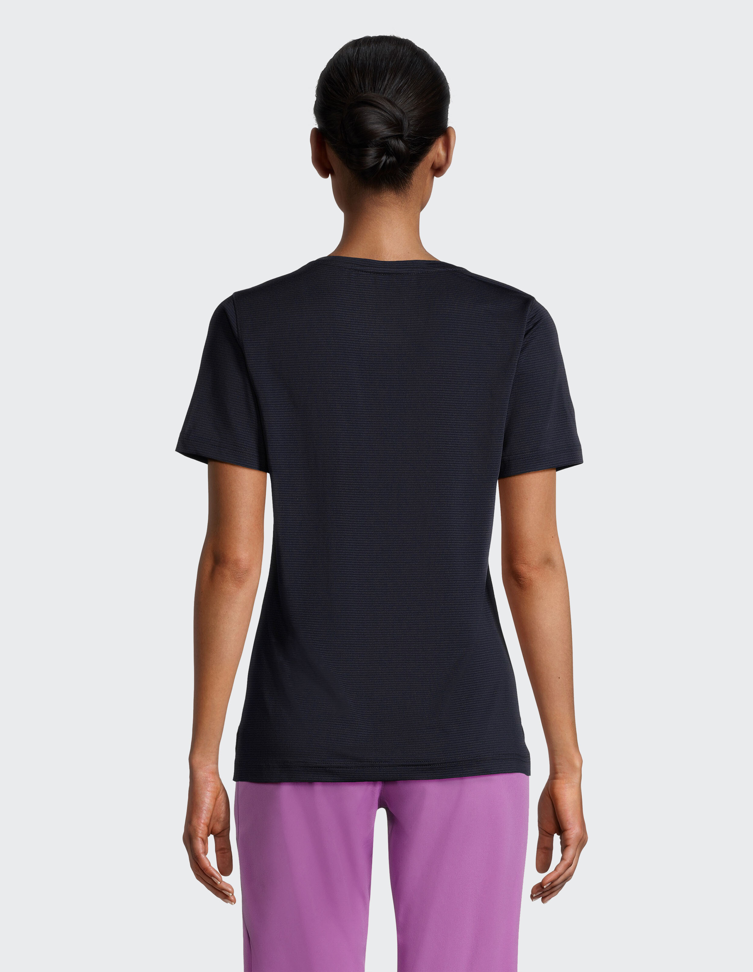 Person von hinten mit dunkelblauem T-Shirt und lila Hose, Haare streng nach hinten gebunden, steht vor hellem Hintergrund.