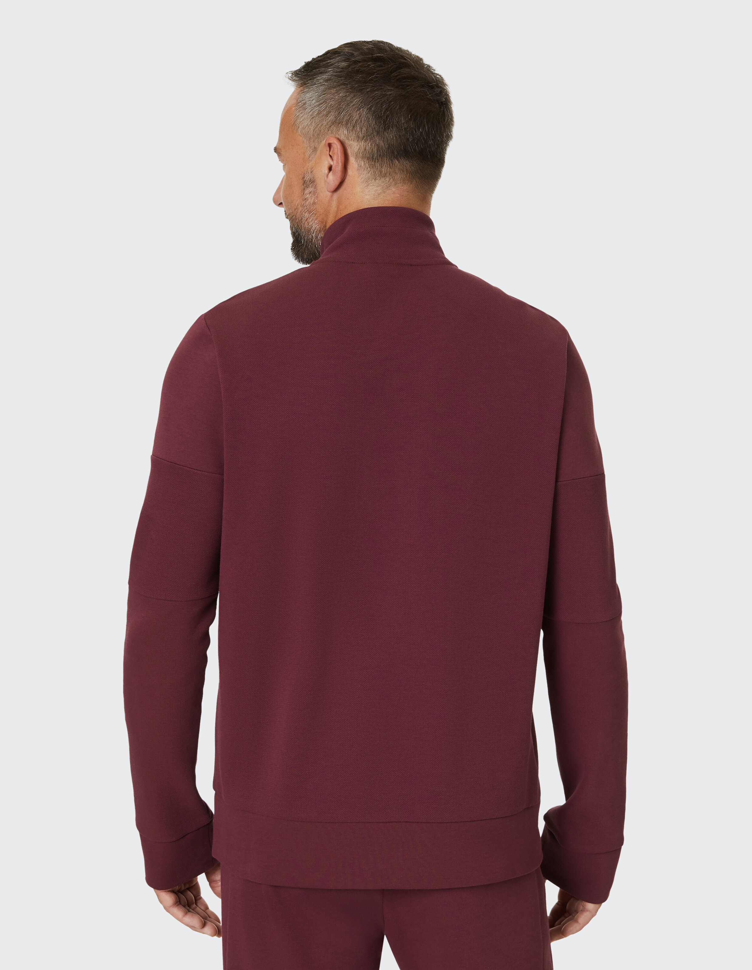 Mann von hinten in bordeauxfarbenem Pullover und passender Hose mit kurzem Haar, stehend vor neutralem Hintergrund.
