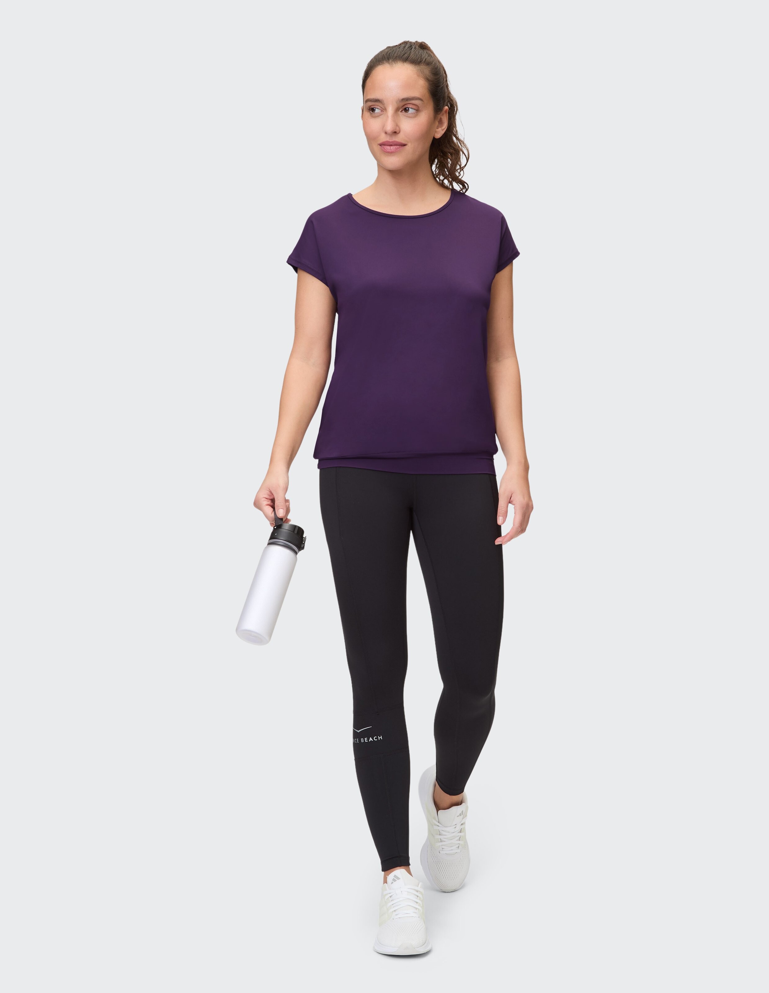 Frau trägt ein lila Freizeit-T-Shirt und schwarze Leggings und hält eine Wasserflasche in der Hand.