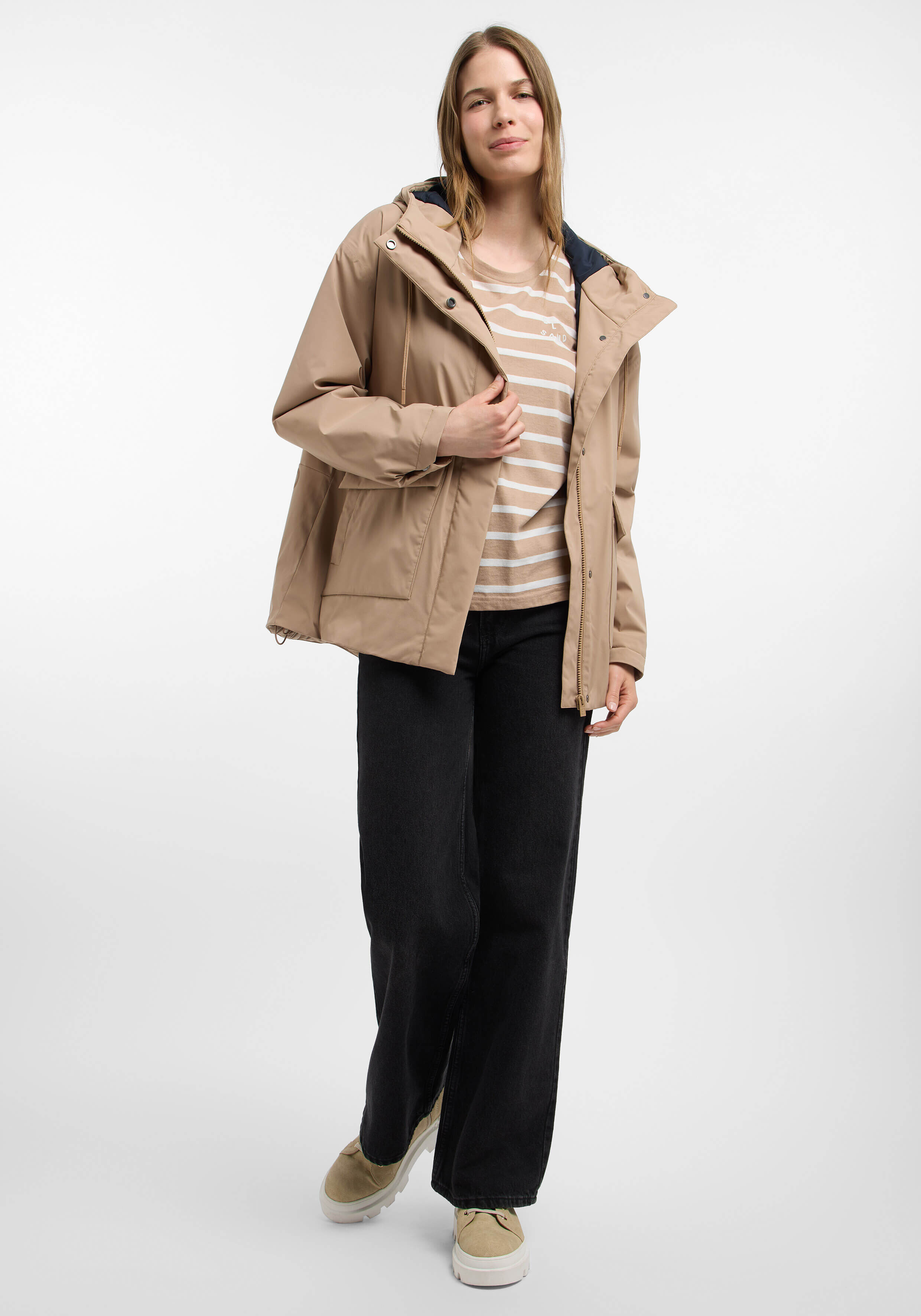 Frau trägt eine beige Kapuzenjacke, ein gestreiftes Hemd und schwarze Jeans für einen lässigen Look.