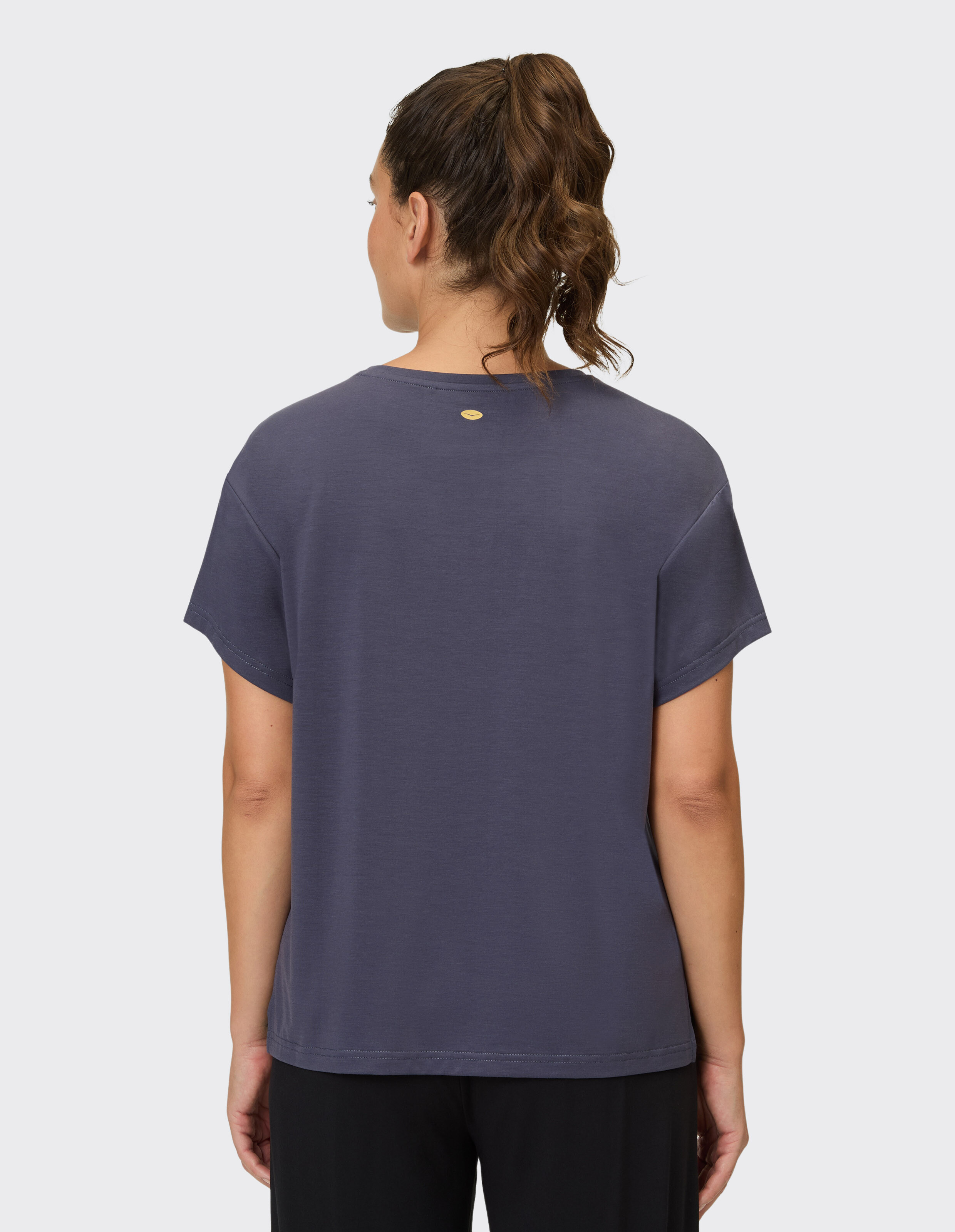 Rückansicht einer Frau, die ein gemütliches dunkelgraues Freizeit-T-Shirt mit einem kleinen goldenen Logo-Detail trägt.