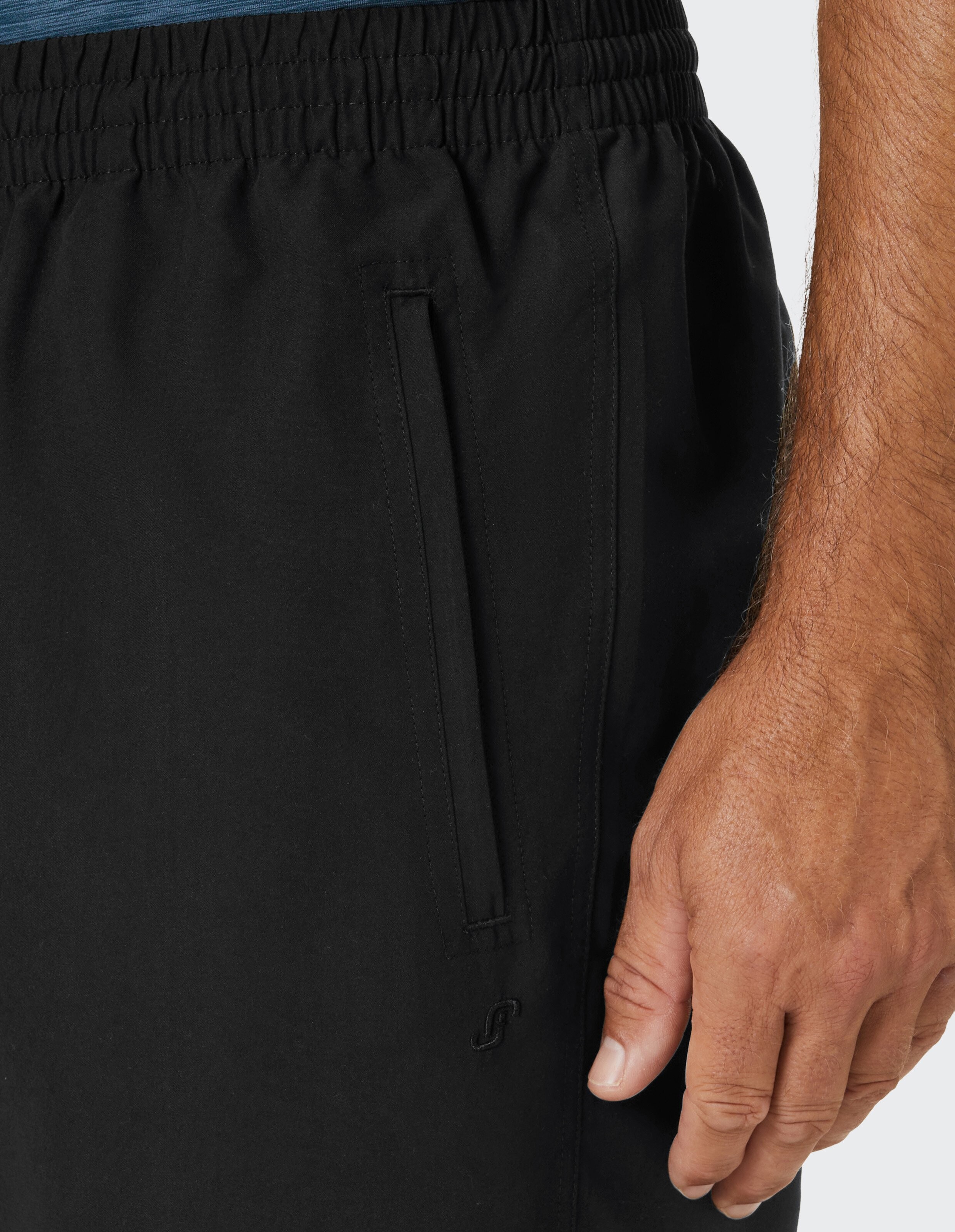 Nahaufnahme einer schwarzen Sporthose mit elastischem Bund und seitlicher Eingrifftasche, daneben eine rechte Männerhand.