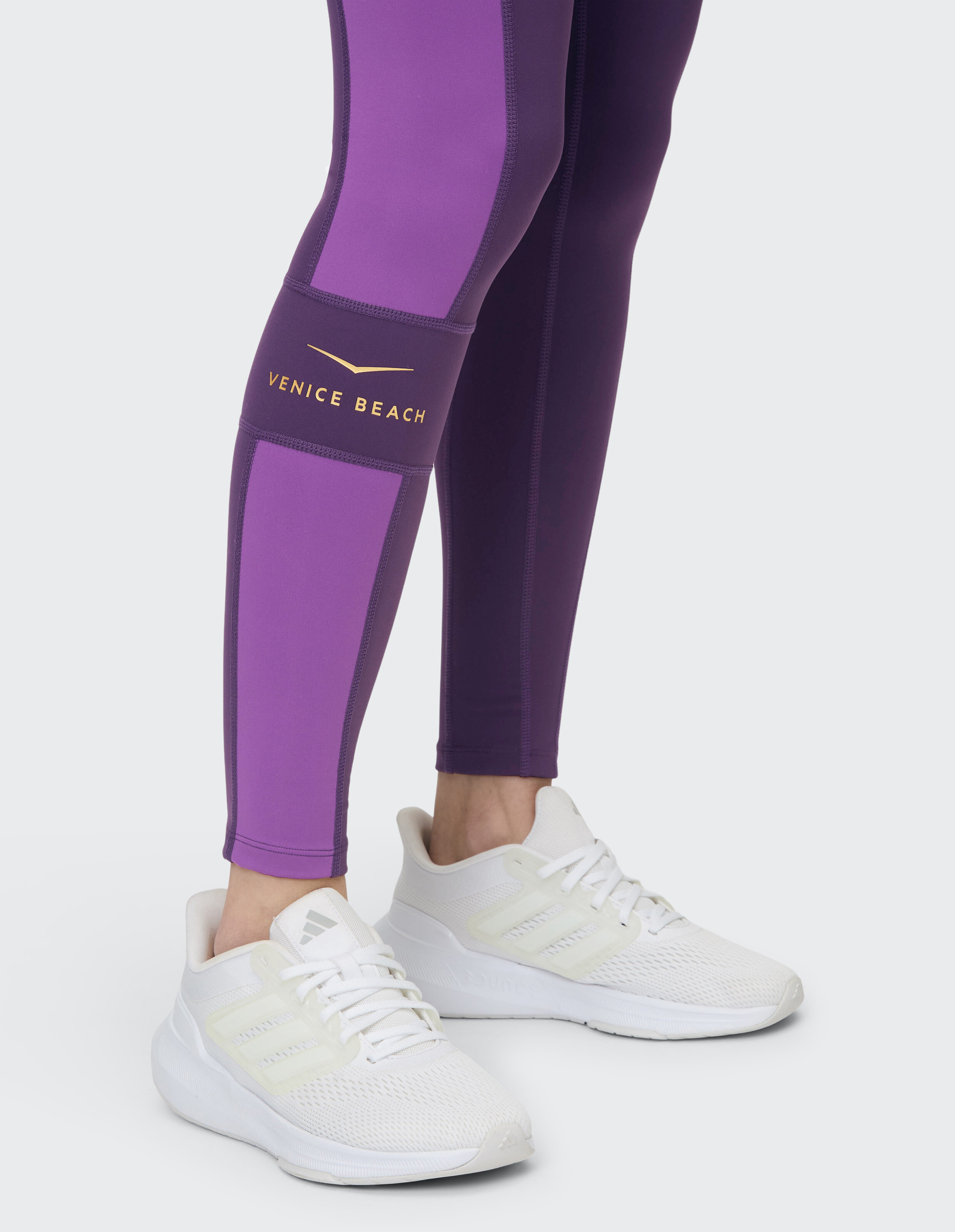 Venice Beach Camy-Strumpfhose in Lila mit goldenem Logo, gepaart mit weißen Sport-Sneakern.