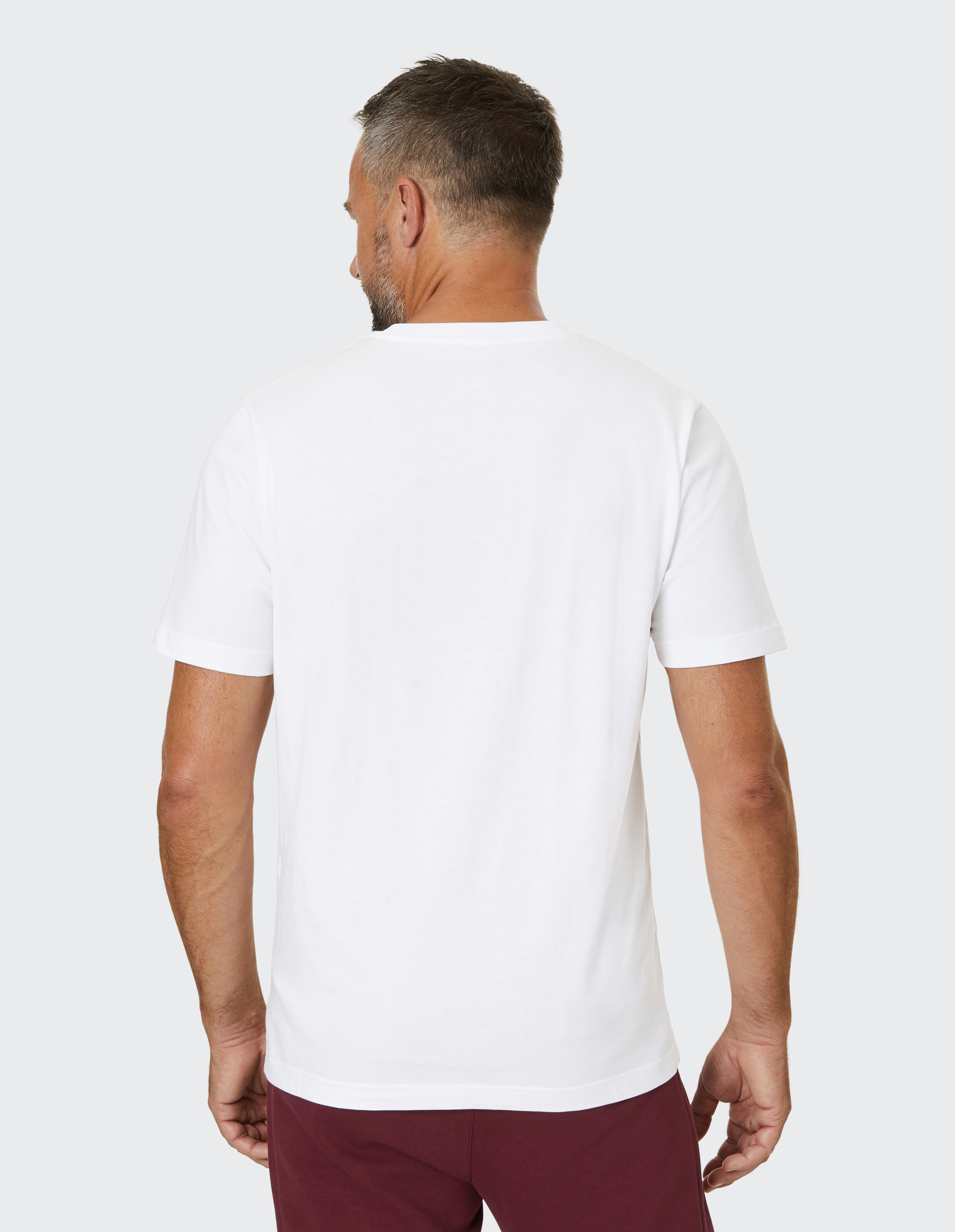 Mann mit Kurzhaarfrisur von hinten in weißem T-Shirt und bordeauxroter Hose vor grauem Hintergrund stehend.