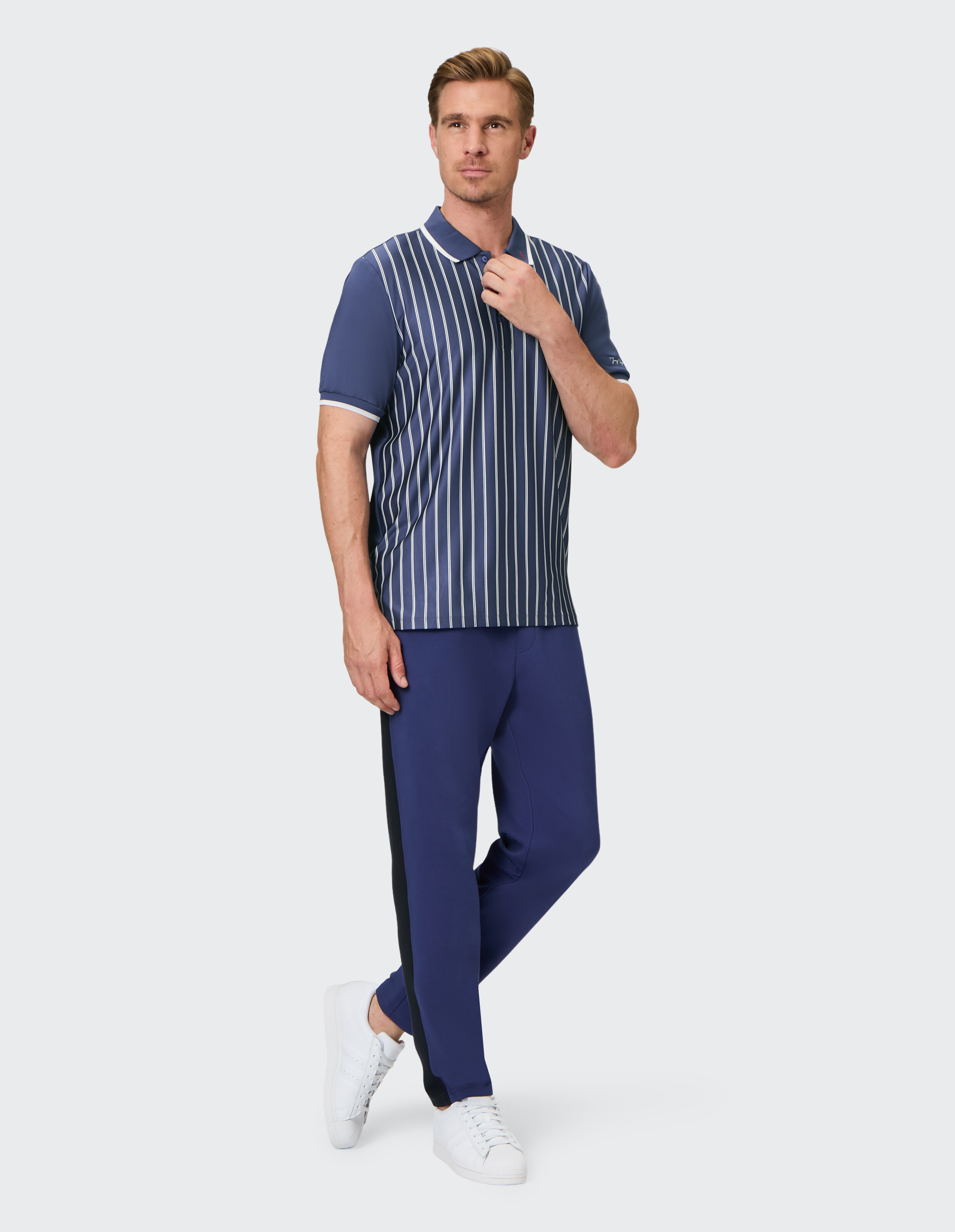 Ein Mann trägt ein blau-weiß gestreiftes Polohemd, blaue Hosen mit schwarzem Streifen und weiße Turnschuhe vor grauem Hintergrund.