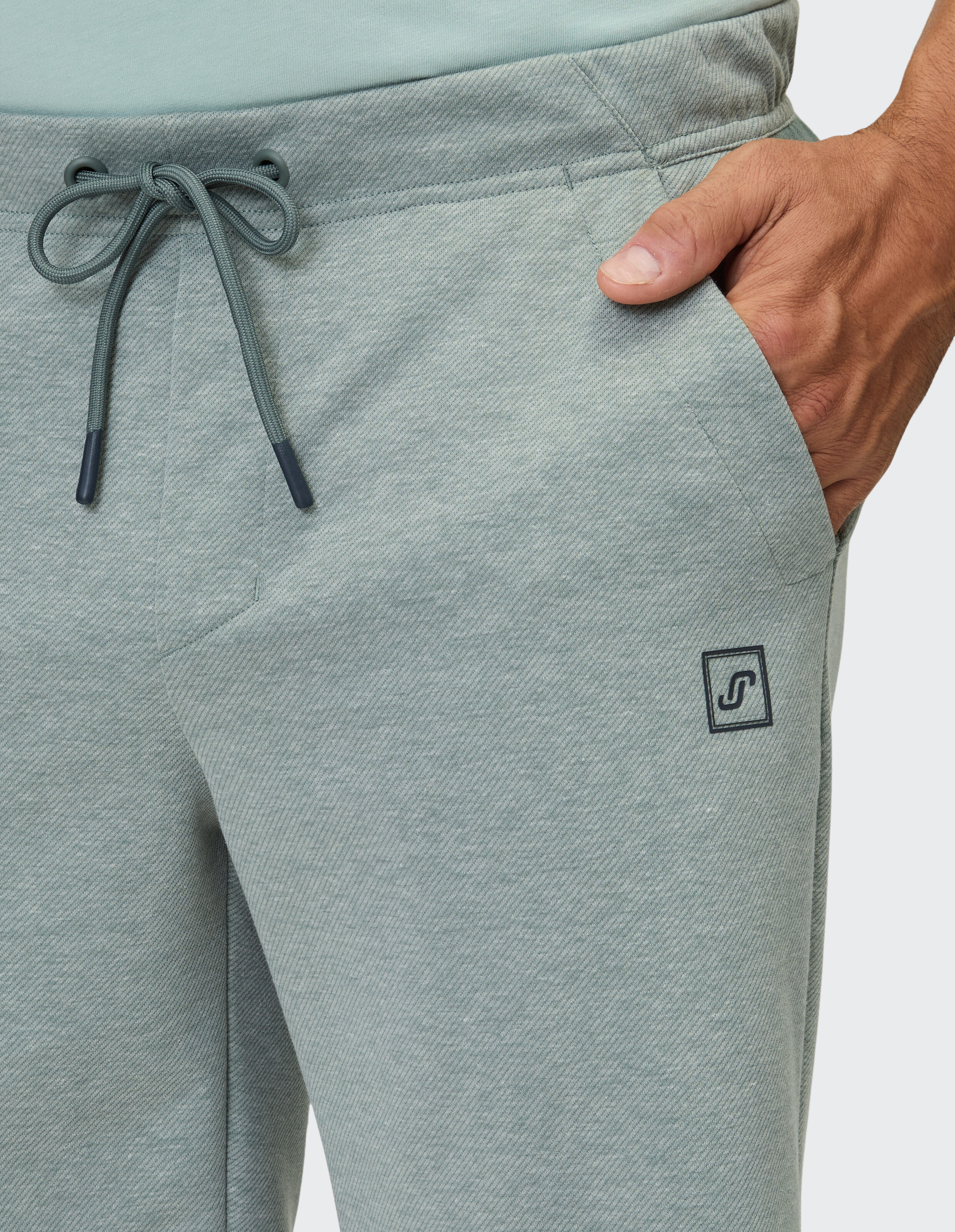 Grüne Jogginghose mit Kordelzug und Logo-Patch, mit seitlicher Hand in der Tasche.
