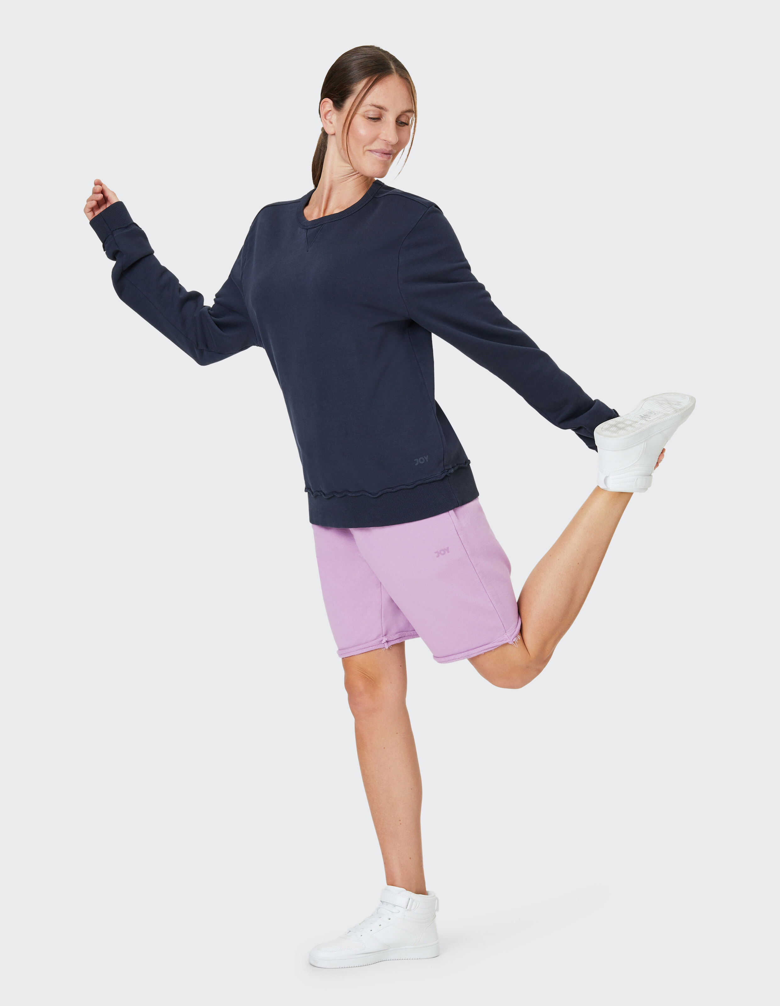 Frau in dunkelblauem Pullover und lila Shorts, steht auf einem Bein, dehnt das andere Bein nach hinten, schaut entspannt.