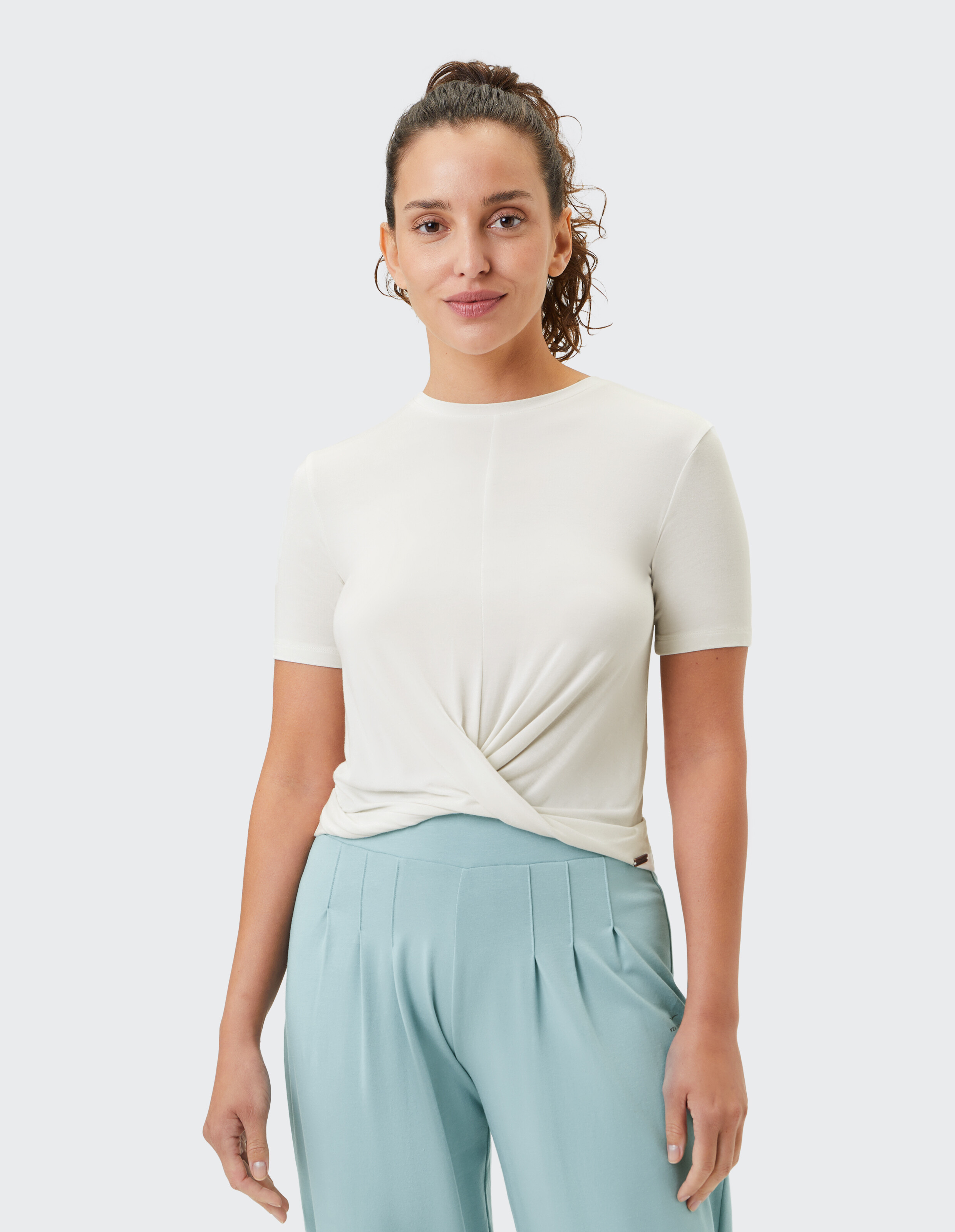 Frau in einem hellcremefarbenen, geknoteten T-Shirt und einer weichen blauen Bundfaltenhose.