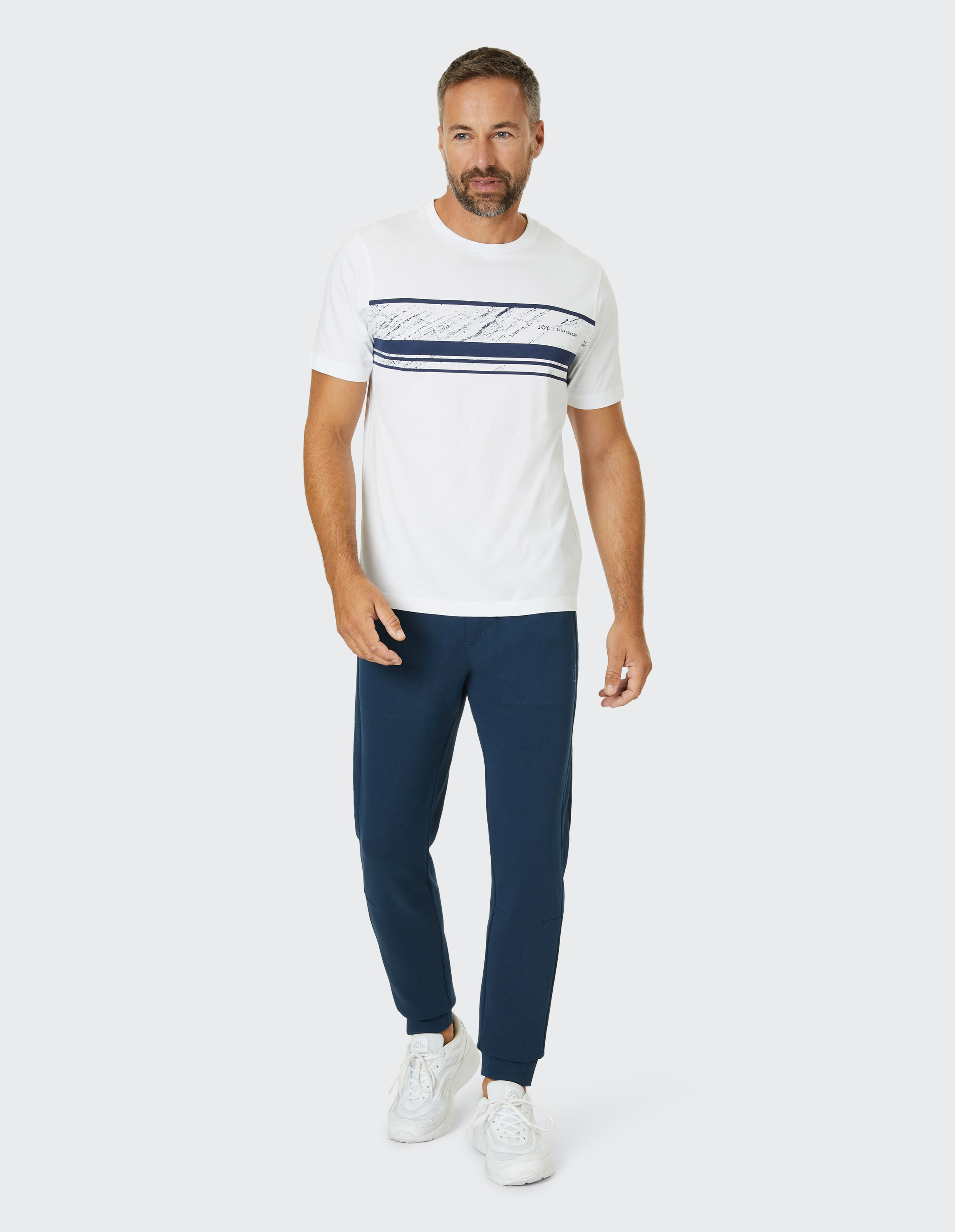Mann in weißem T-Shirt mit blauen Streifen, blauer Jogginghose und weißen Sneakers vor grauem Hintergrund gehend.