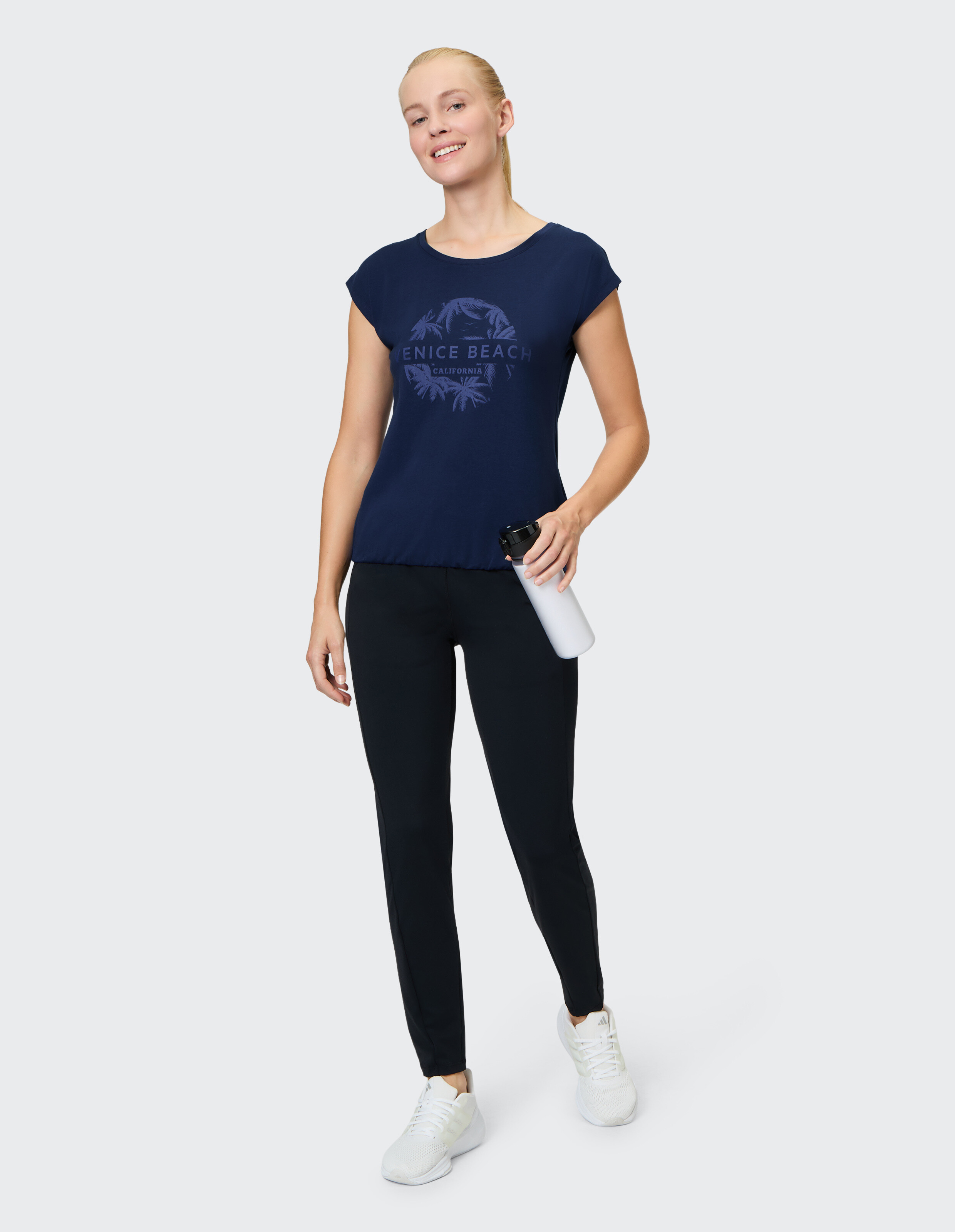 Frau in marineblauem Grafik-T-Shirt und schwarzen Leggings, eine Wasserflasche in der Hand, lässiger Stil.