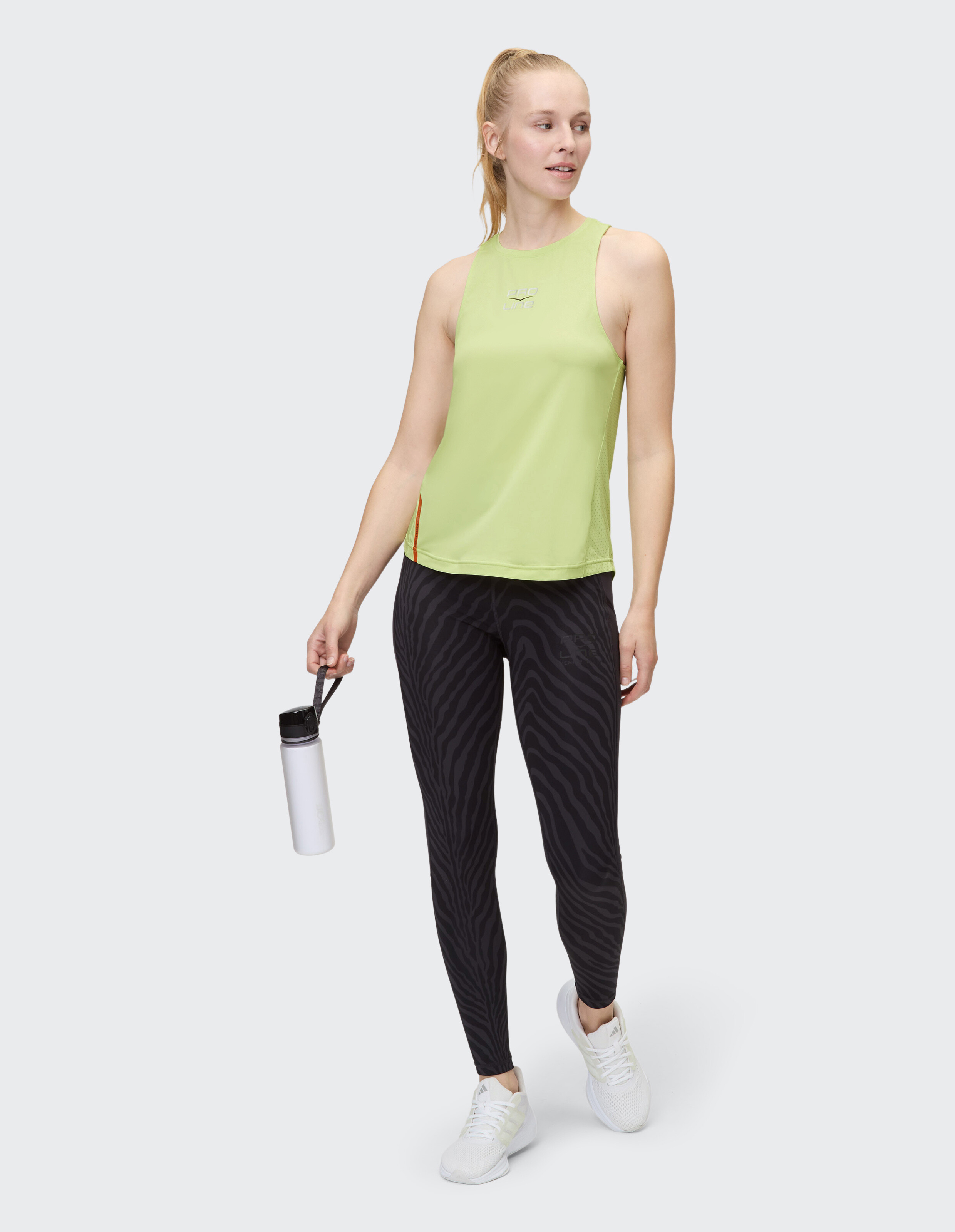Frau in einem hellgrünen Tanktop und schwarzen Leggings mit Zebramuster, hält eine Wasserflasche in der Hand.