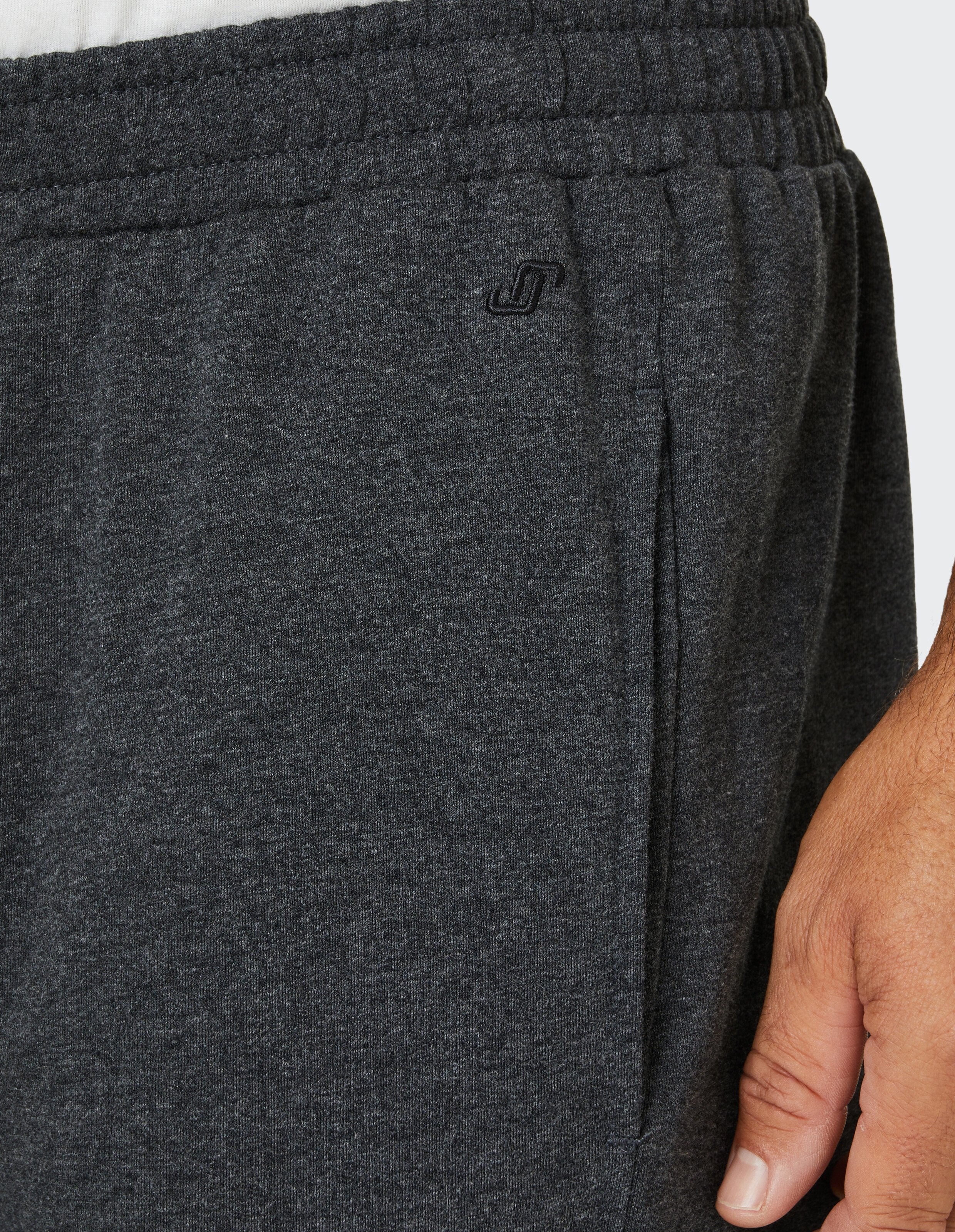 Nahaufnahme einer dunkelgrauen Jogginghose mit elastischem Bund, einer Handtasche und einem kleinen Logo, getragen von einer männlichen Hand.