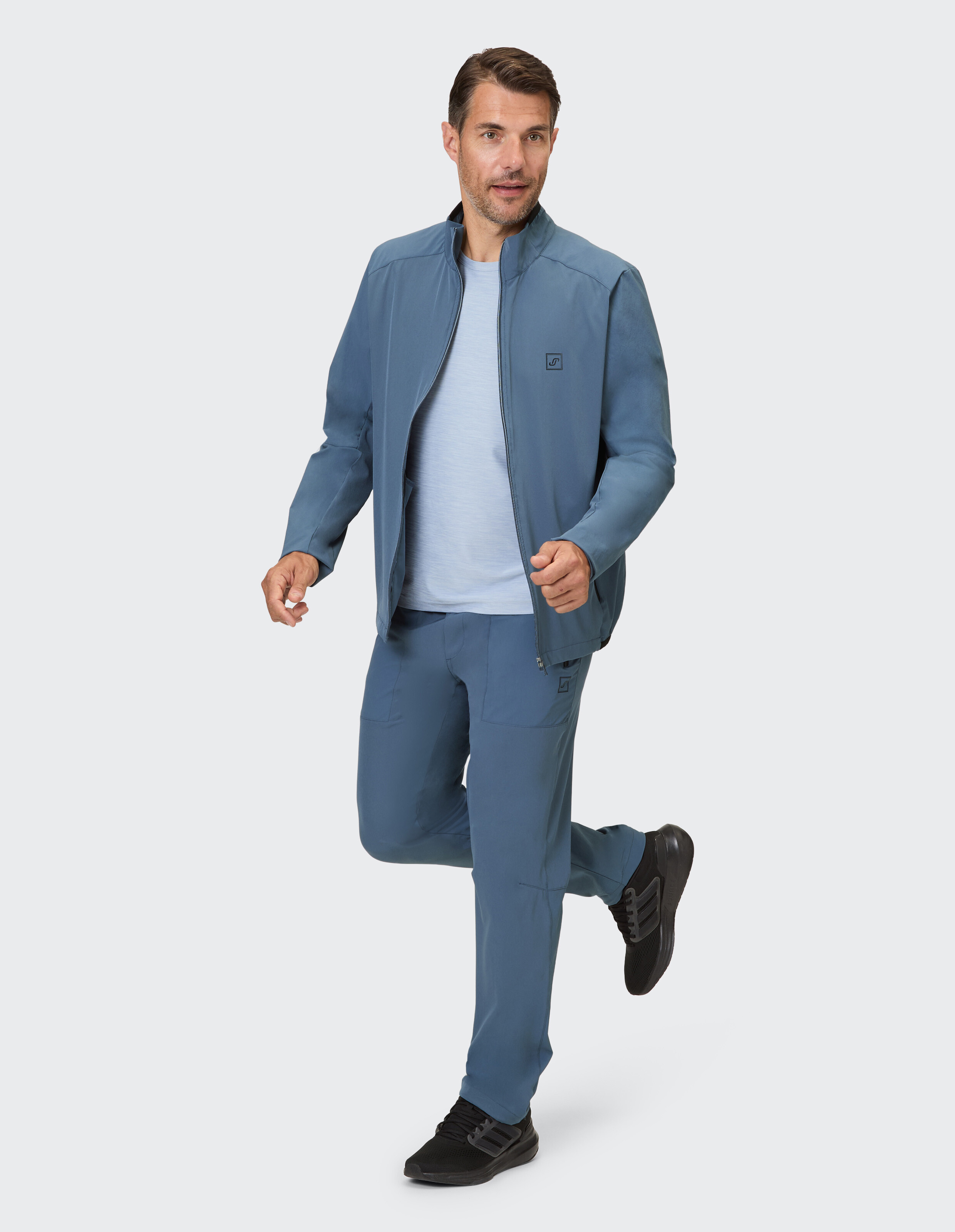 Ein Mann in einem blauen Trainingsanzug mit durchgehendem Reißverschluss und Hose, lässig auf einem Bein stehend.