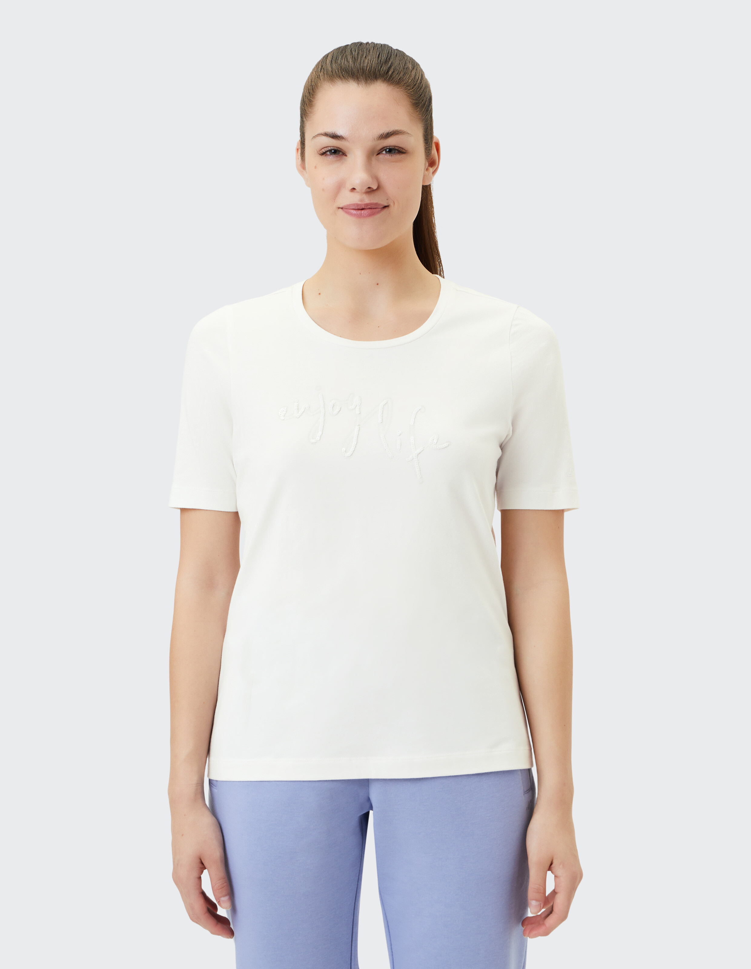 Frau trägt ein weißes T-Shirt mit „Enjoy Life“-Stickerei, gepaart mit einer hellblauen Hose.