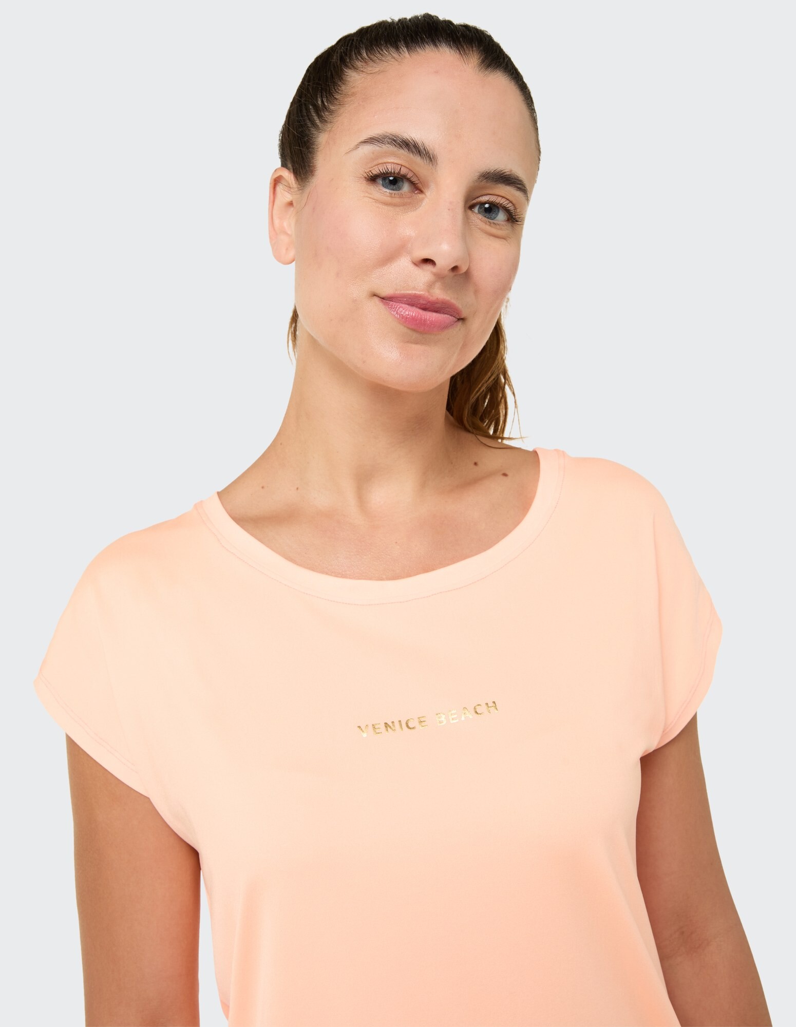 Nahaufnahme einer Frau in einem pfirsichfarbenen T-Shirt mit „VENICE BEACH“-Aufdruck im lässigen Stil.