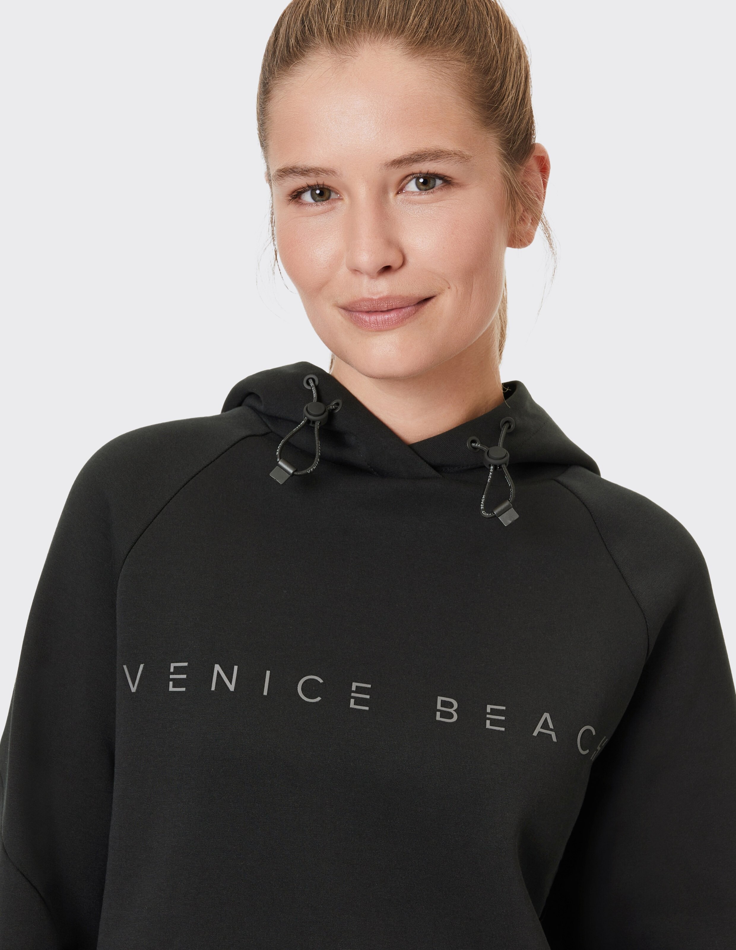 Frau trägt einen schwarzen Leny-Hoodie von Venice Beach mit Kapuze mit Kordelzug.