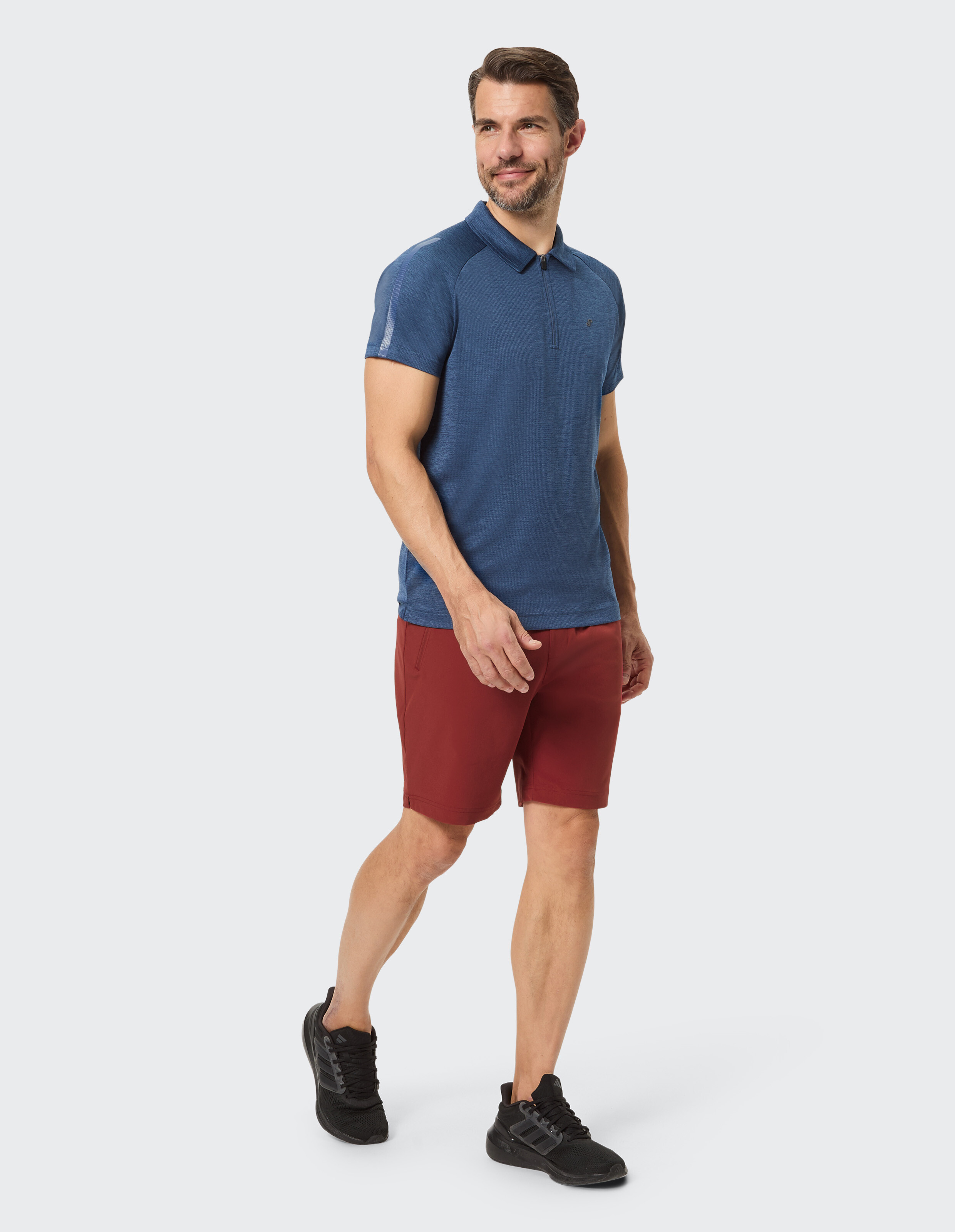 Ein Mann steht in sportlicher Kleidung mit blauem Poloshirt, roten Shorts und schwarzen Sneakers vor einem weißen Hintergrund.
