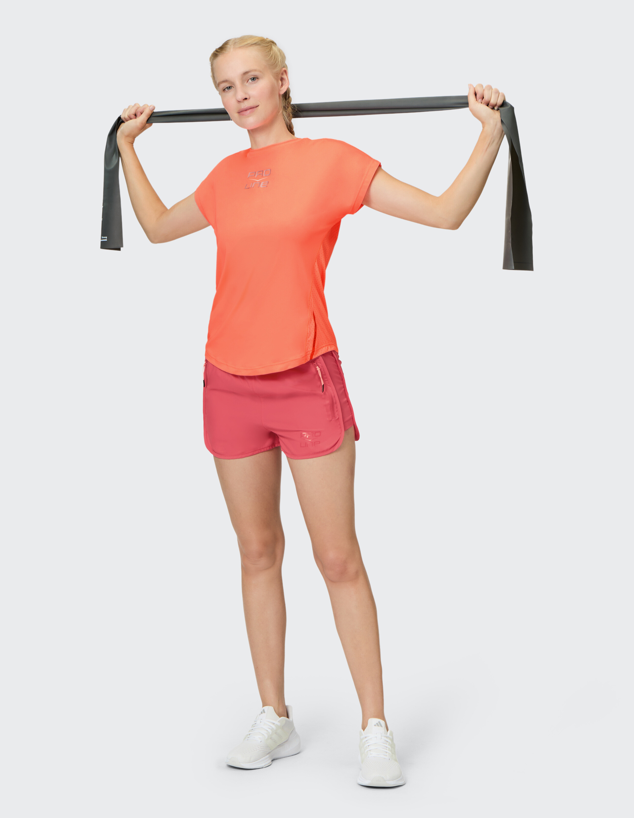 Frau in korallenrotem Sport-T-Shirt und rosa Shorts trainiert mit einem Widerstandsband.