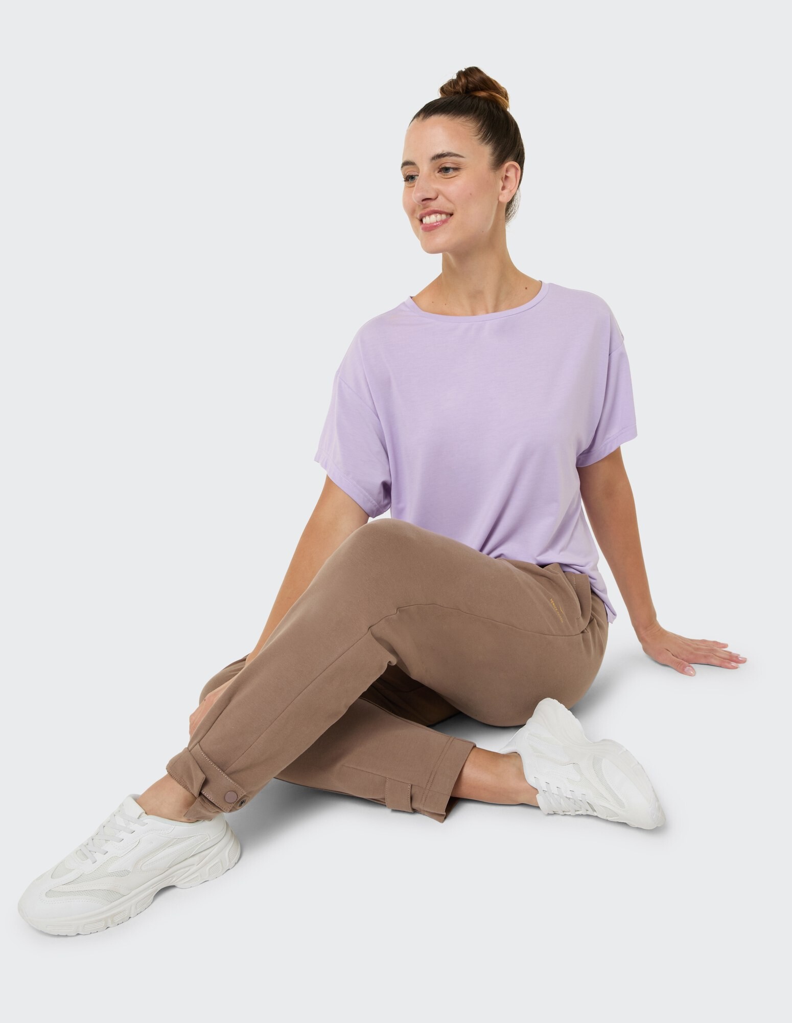 Frau in lavendelfarbenem T-Shirt und brauner Hose sitzt lässig in weißen Turnschuhen.