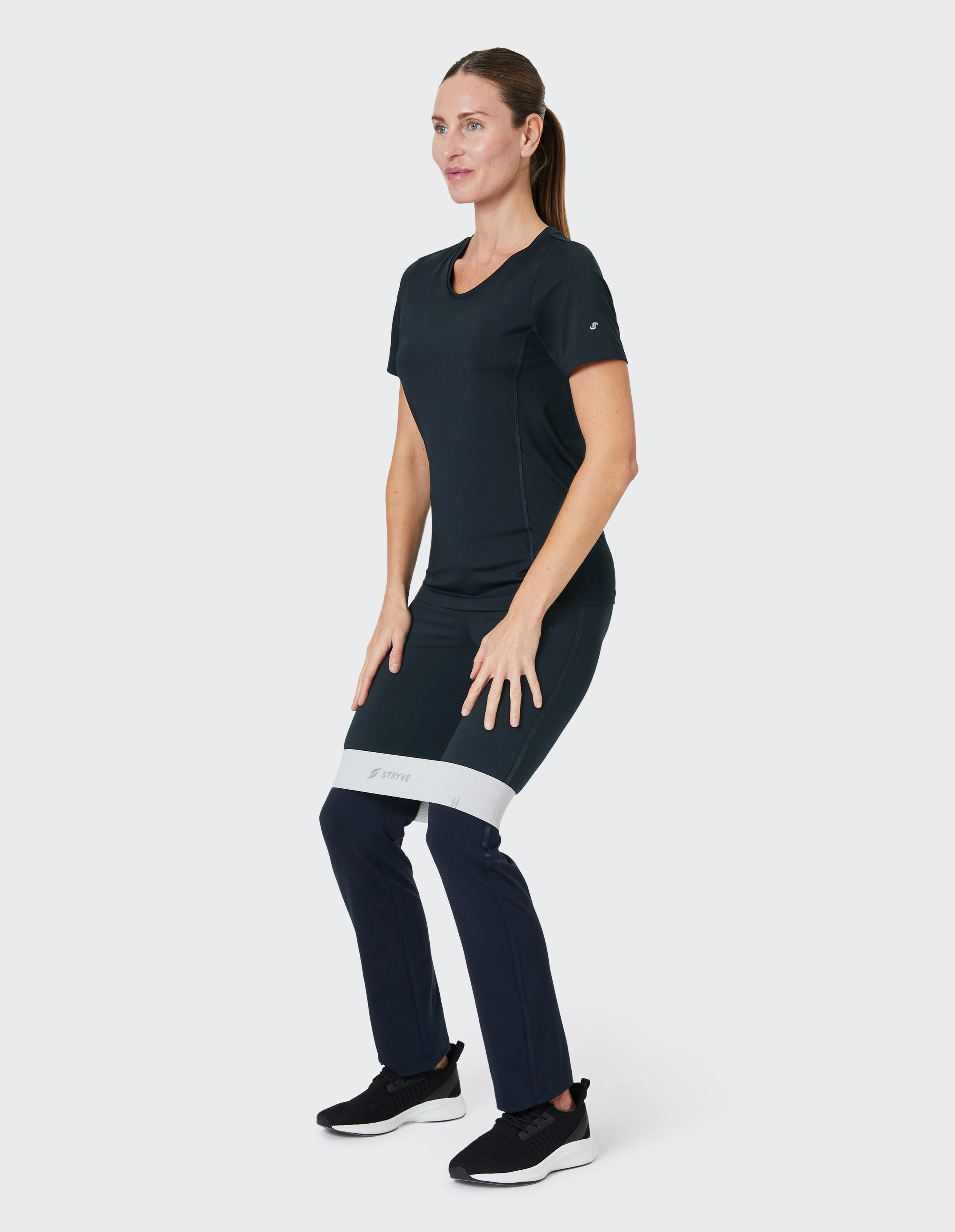 Frau in schwarzer Sportkleidung macht Kniebeuge mit Fitnessband um Oberschenkel, schaut konzentriert zur Seite, auf hellem Hintergrund.