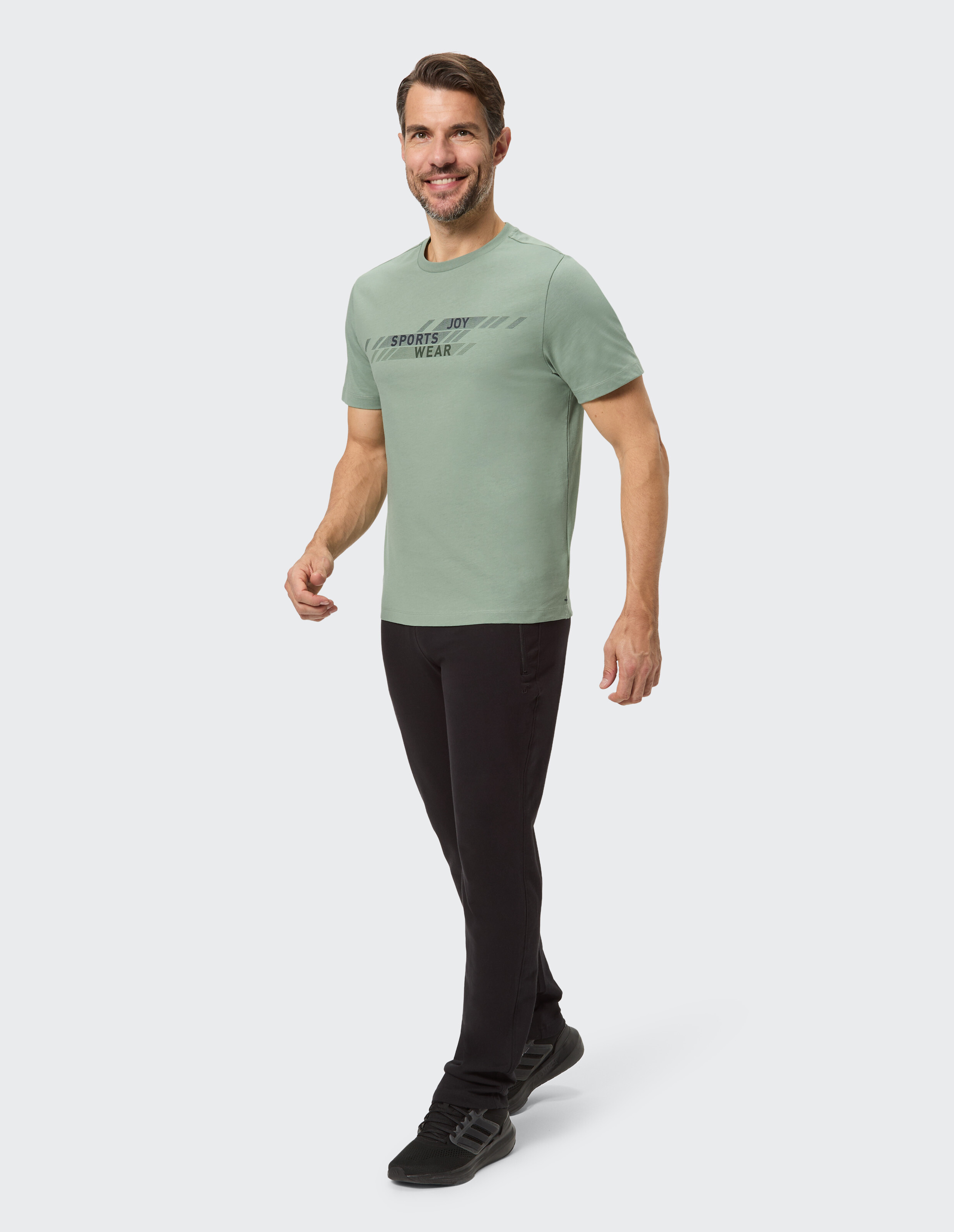 Mann in hellgrünem Sport-T-Shirt und schwarzer Hose, lässig lächelnd.