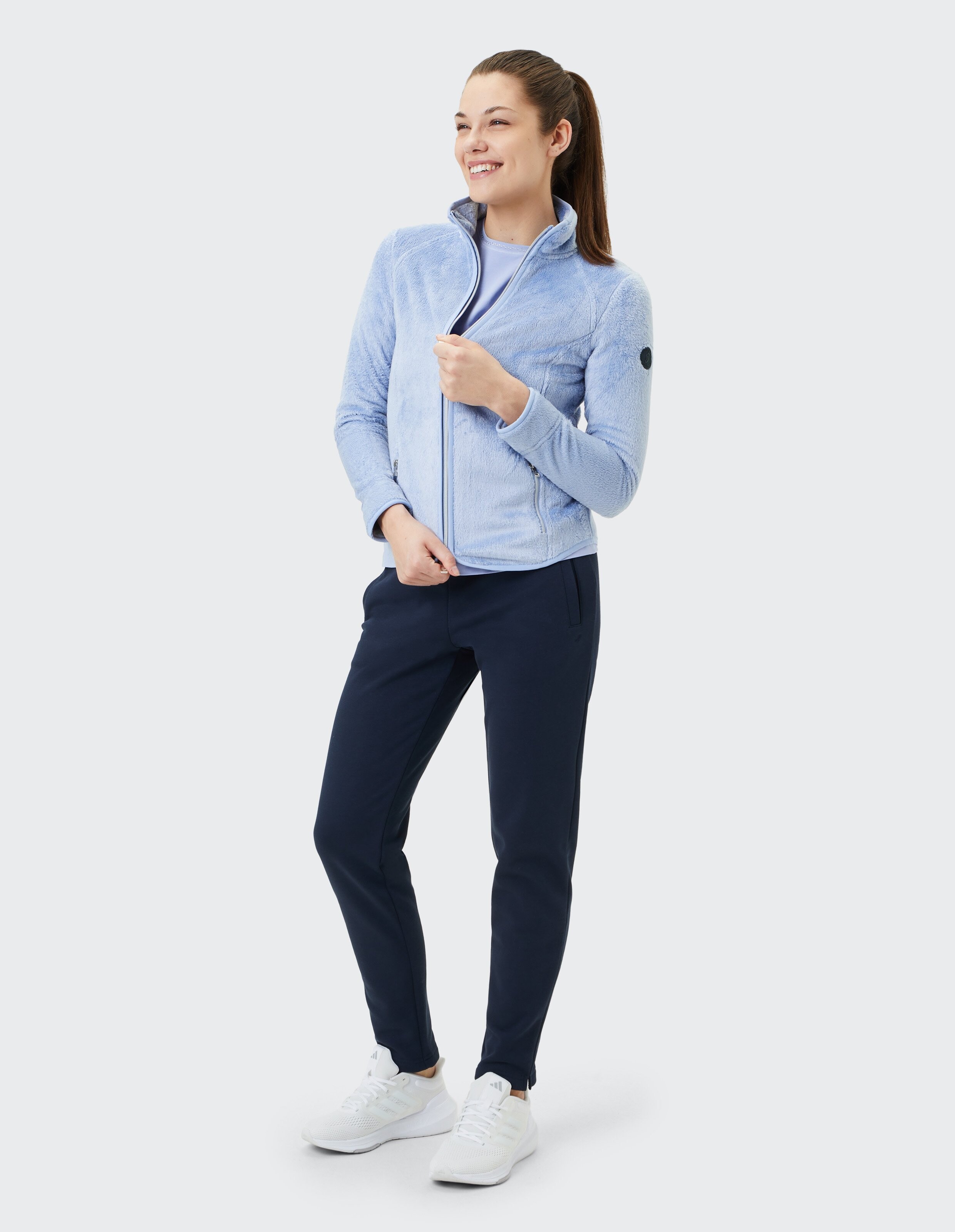 Frau in hellblauer Fleecejacke und dunkelblauer Hose, lässig lächelnd.