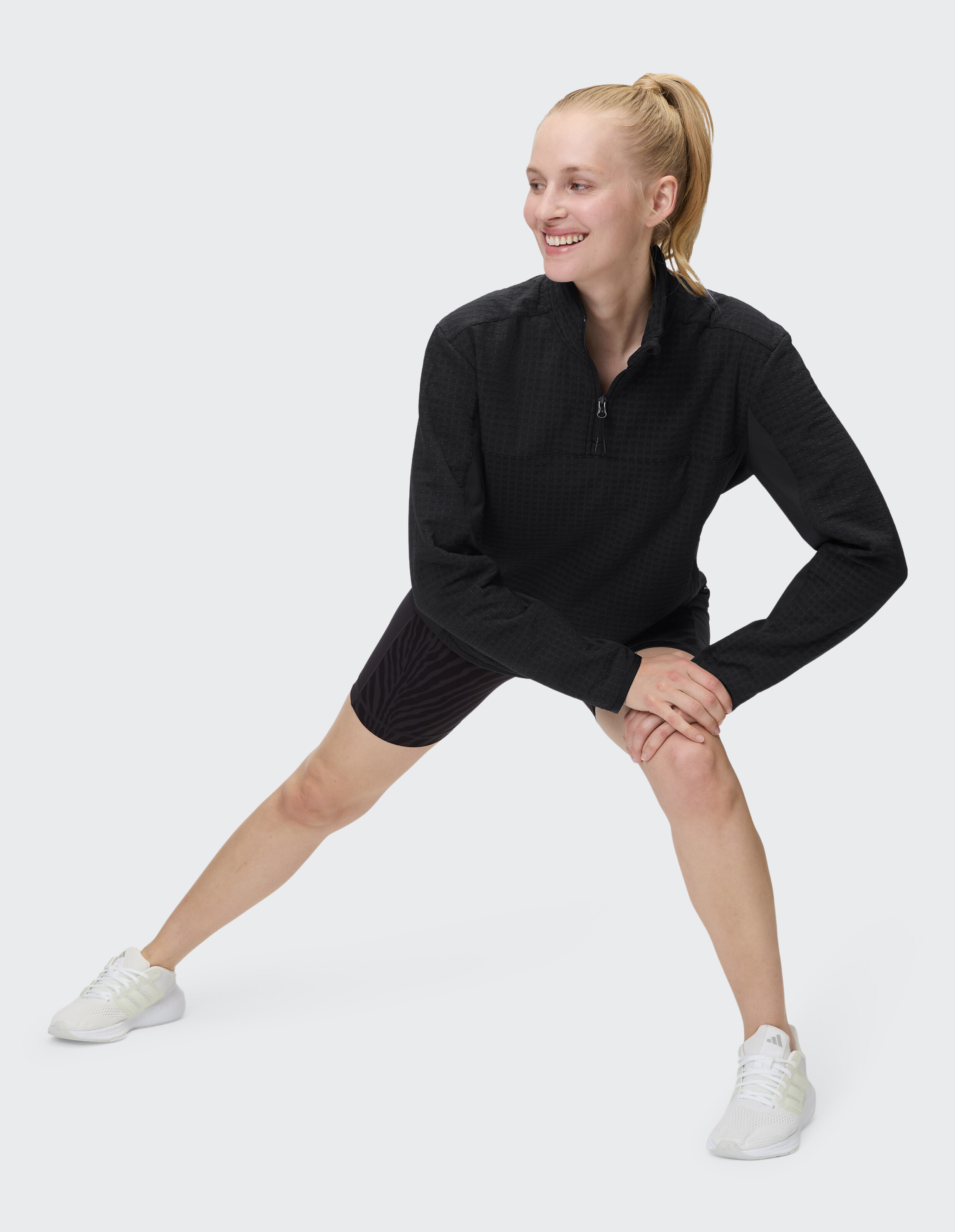 Frau beim Dehnen in einem schwarzen Oberteil mit Reißverschluss und gemusterten Shorts, ideal für Freizeitkleidung.