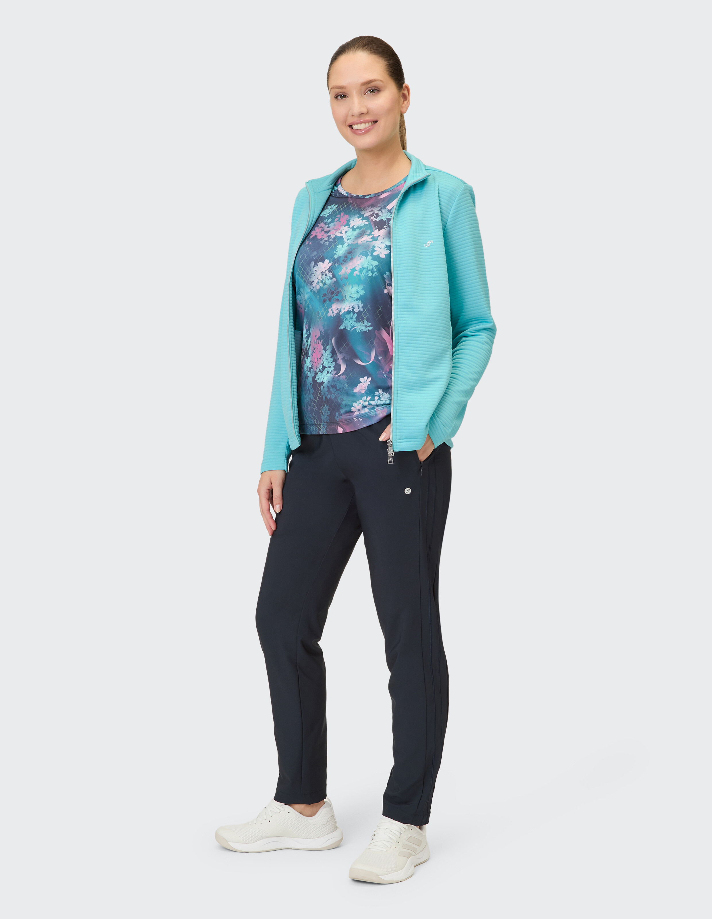 Frau in türkisfarbener Jacke und geblümtem Oberteil, gepaart mit dunkelblauen Hosen und weißen Turnschuhen.